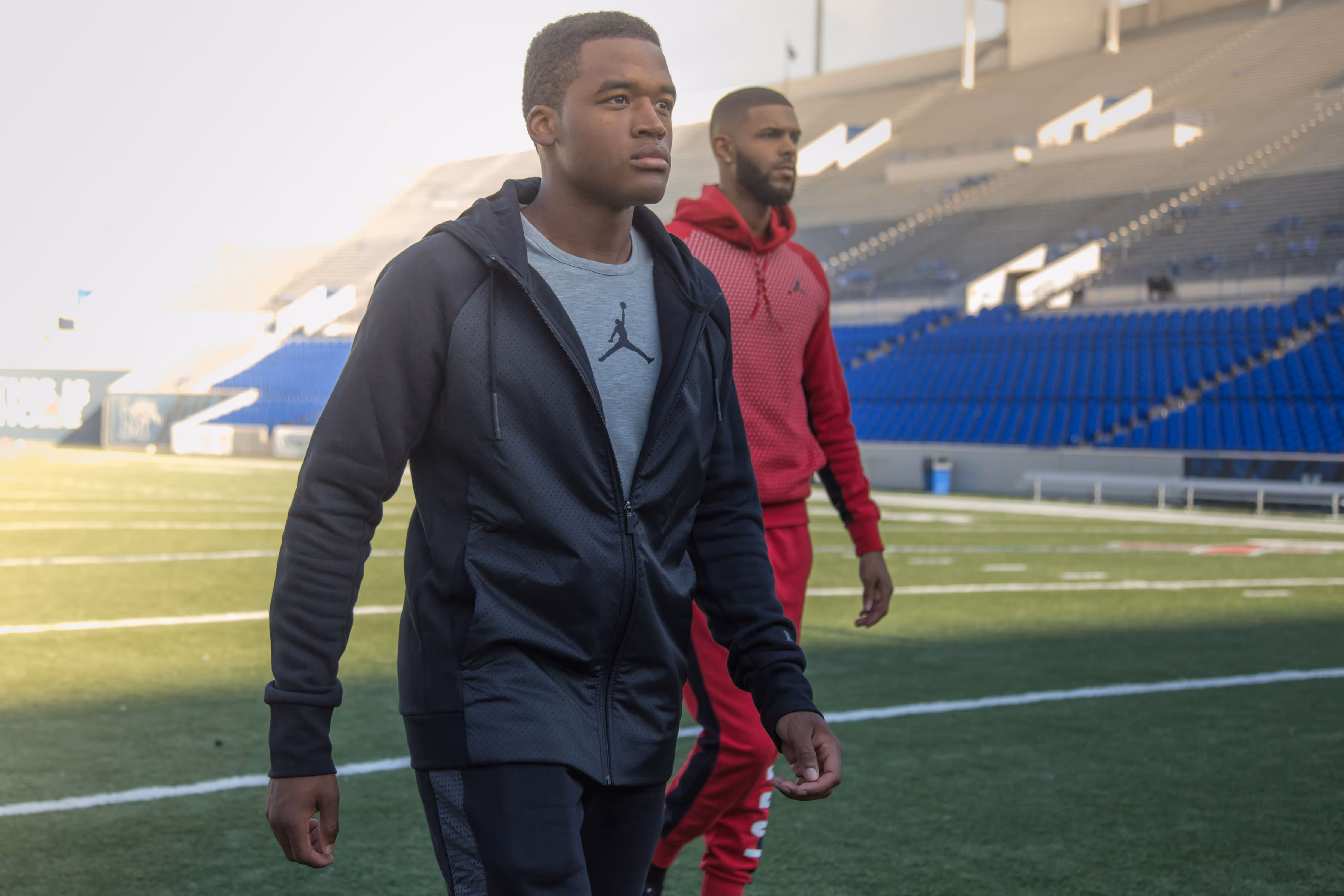 Two men walking on a football field with empty blue stadium seats in the background, one in black athletic wear and the other in red.