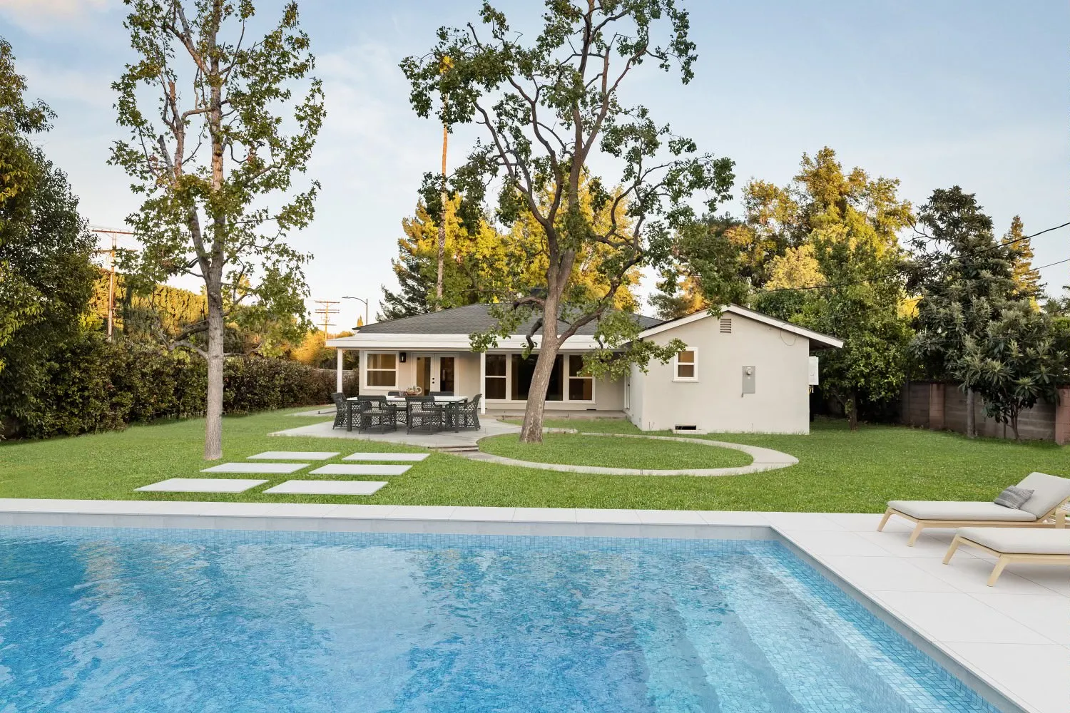 Backyard view of a single-story house with a patio table and chairs, green lawn, trees, and a blue swimming pool with lounge chairs.