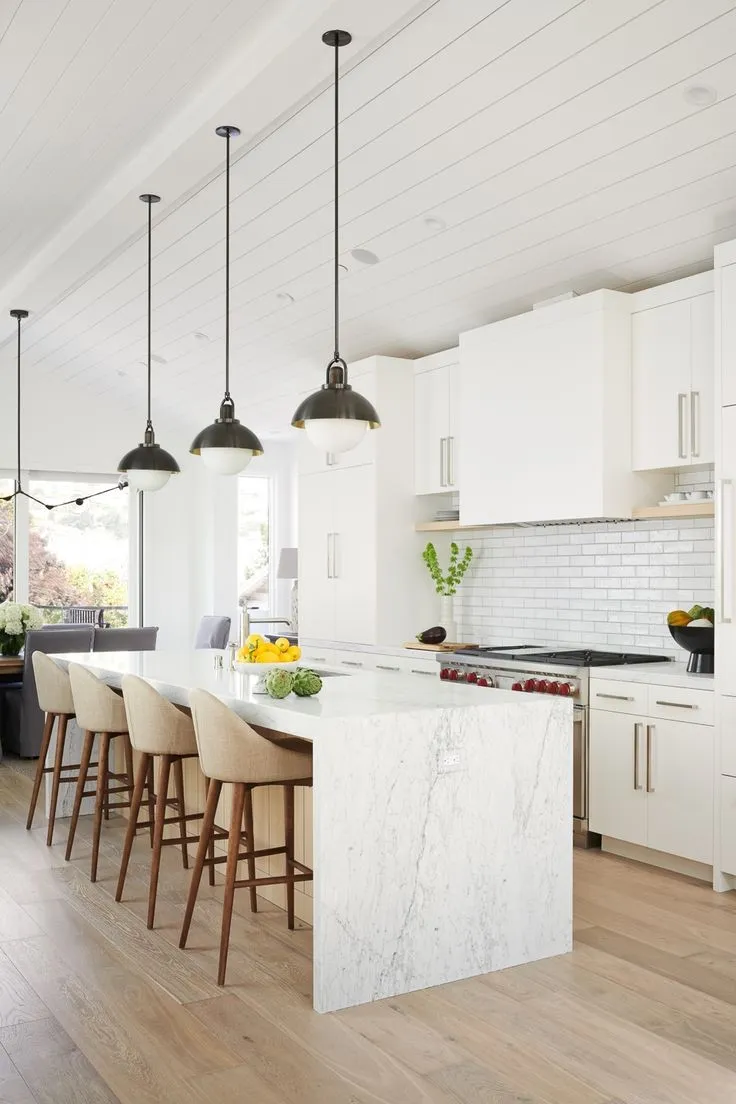 Modern kitchen with white marble waterfall island, beige bar stools, pendant lights, and white cabinetry with subway tile backsplash.