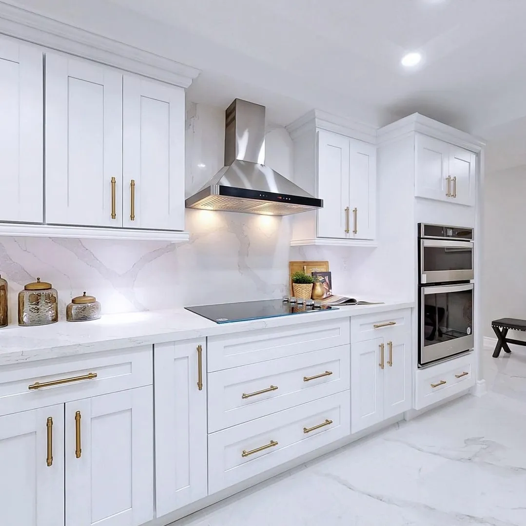 Modern white kitchen with shaker cabinets, brass hardware, quartz countertops, stainless steel range hood, and built-in double oven.
