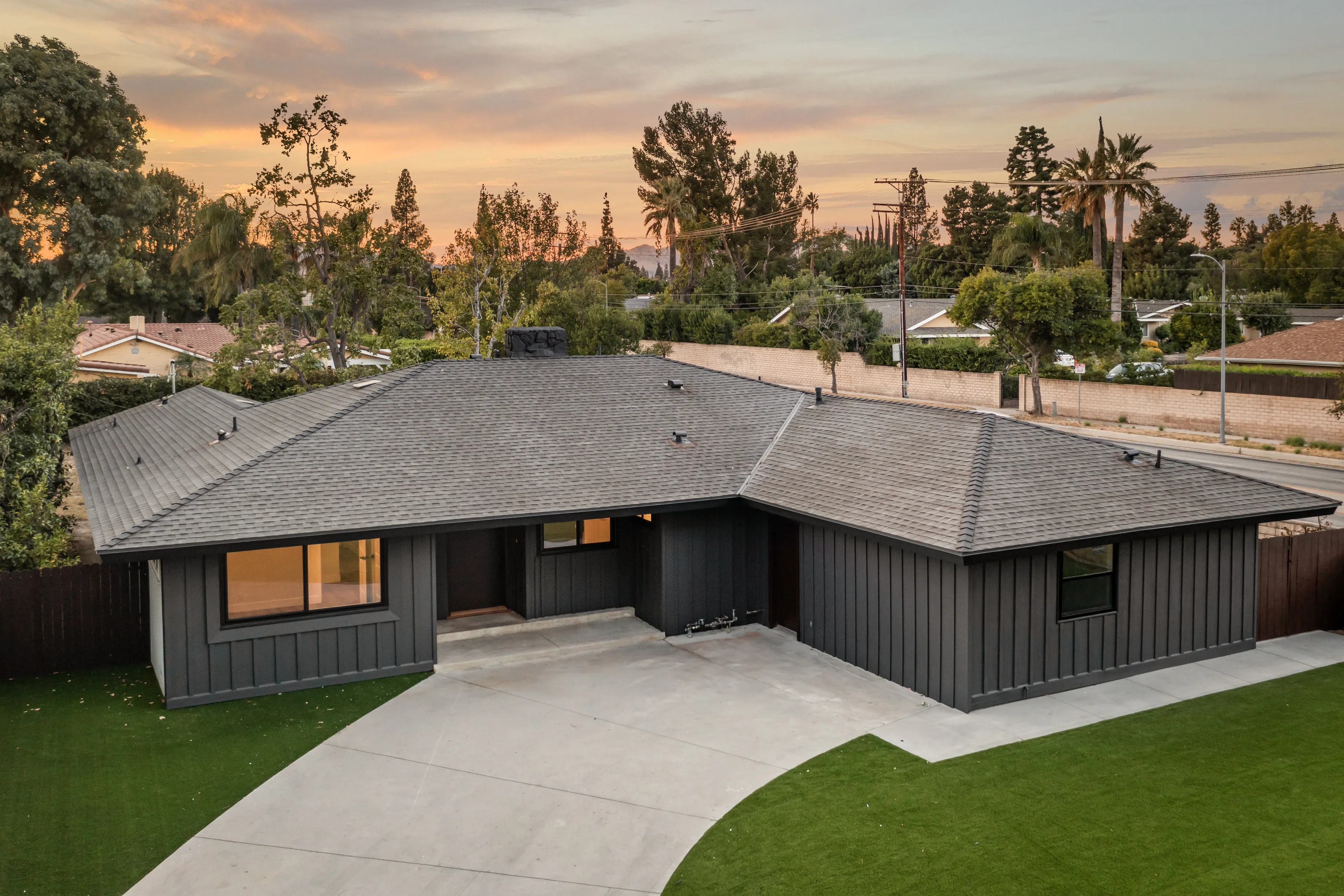 Single-story modern ranch-style house with dark board and batten siding, gray shingled roof, and a wide concrete driveway at sunset.