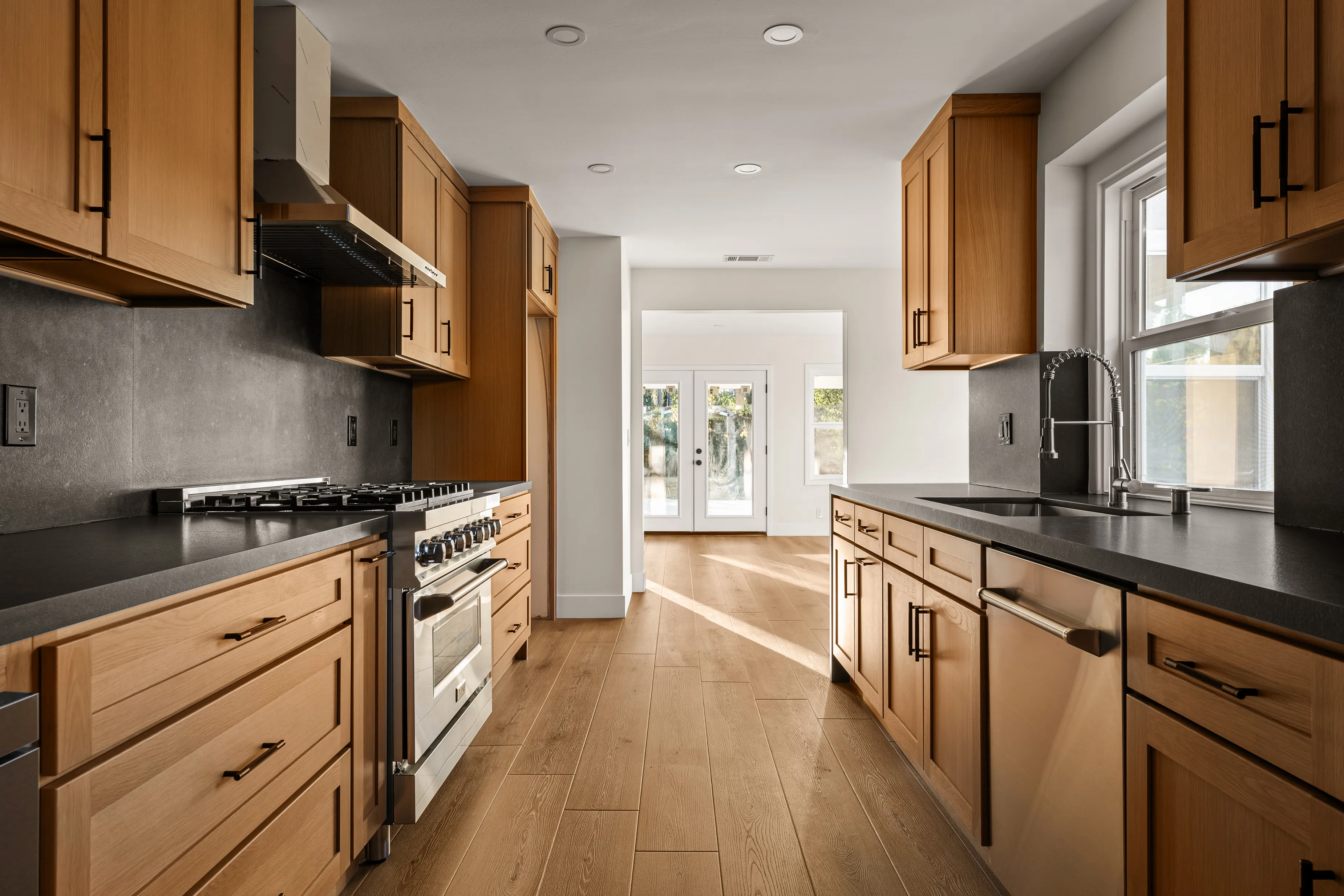Modern kitchen with wooden cabinets, black countertops, stainless steel stove and dishwasher, and wood flooring.