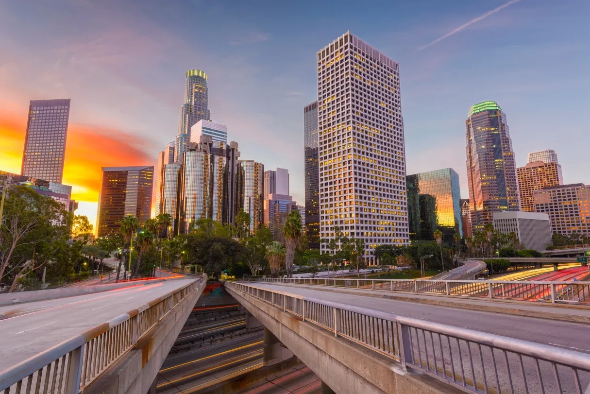 View of downtown Los Angeles skyline at sunset with empty freeway overpasses and palm trees.