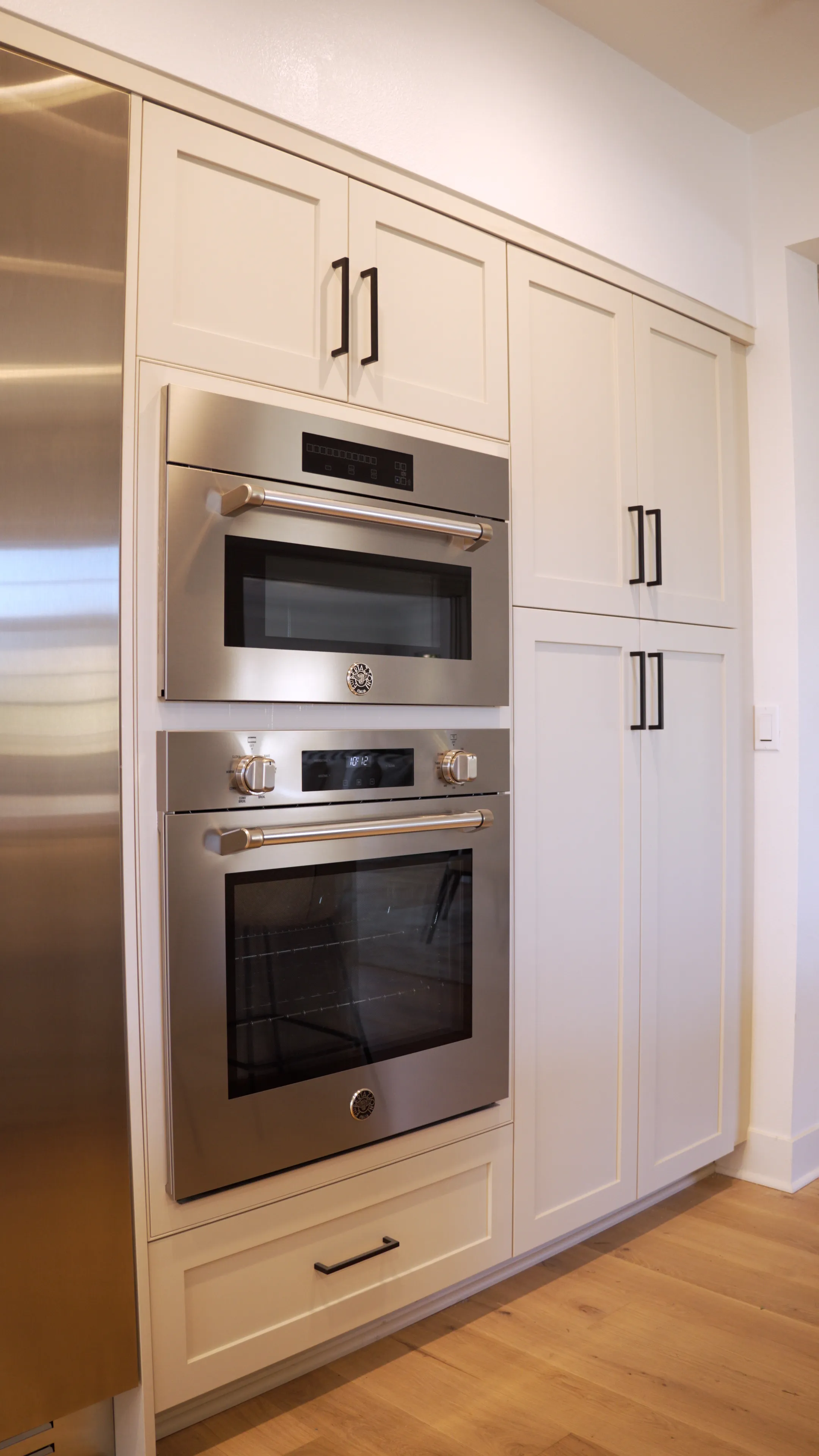 Built-in stainless steel microwave and oven in white kitchen cabinets with black handles on light wood flooring.
