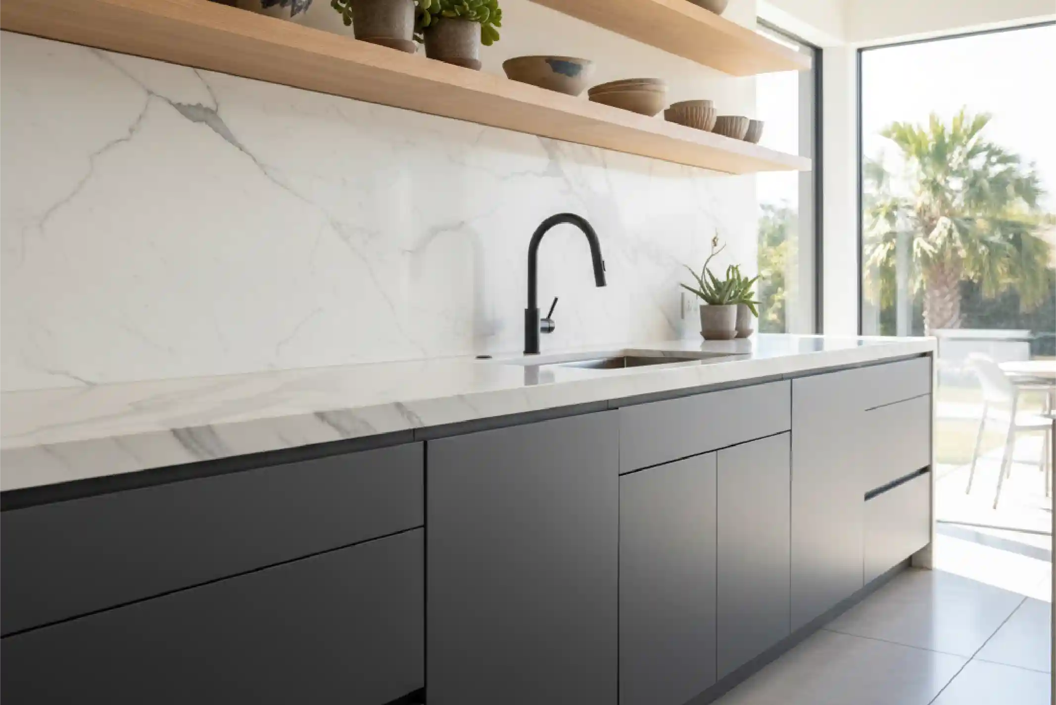 Modern kitchen with dark cabinets, white marble countertop and backsplash, black faucet, open shelves with pottery, and large window showing palm trees.