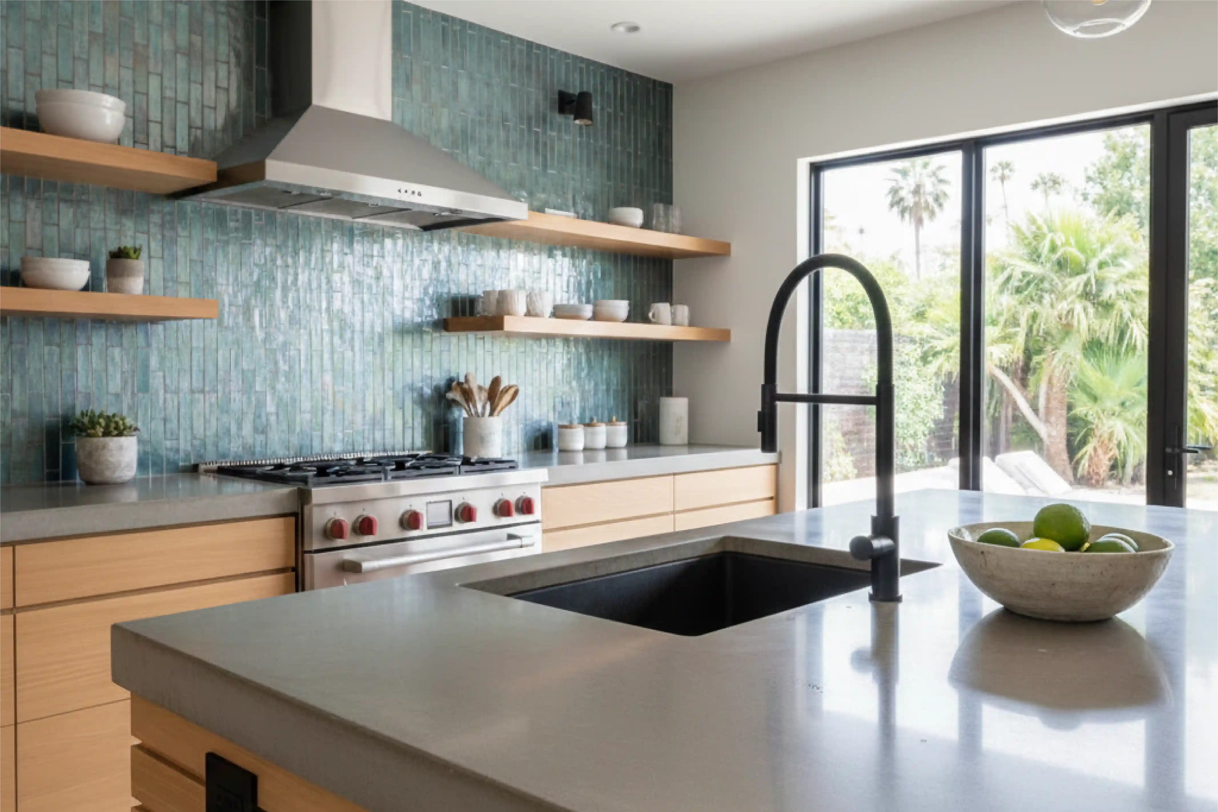 Modern kitchen with light wood cabinets, green subway tile backsplash, stainless steel stove and hood, and large island with black sink and faucet overlooking a garden through glass doors.