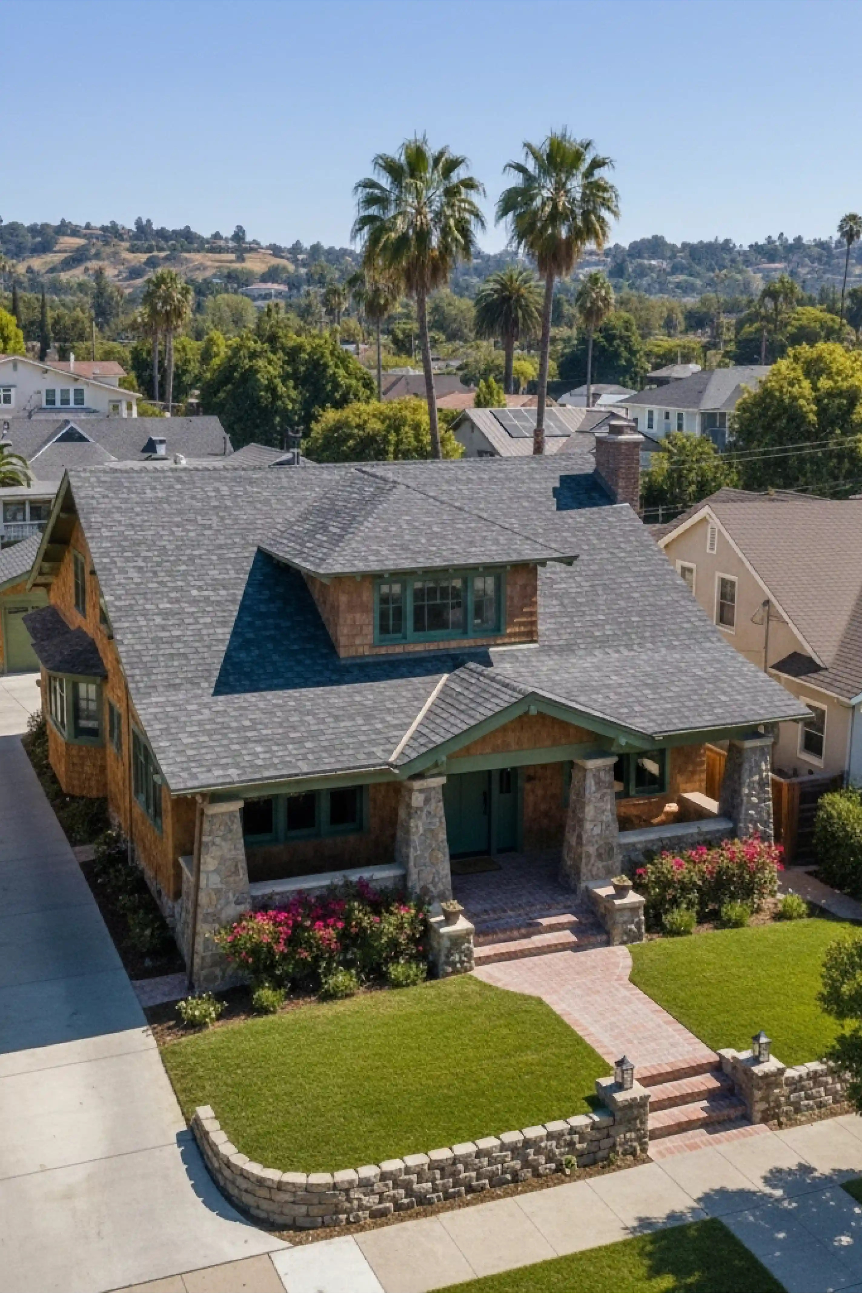 Craftsman-style house with gray shingled roof, stone pillars, green-trimmed windows, and a well-maintained lawn with flower beds.