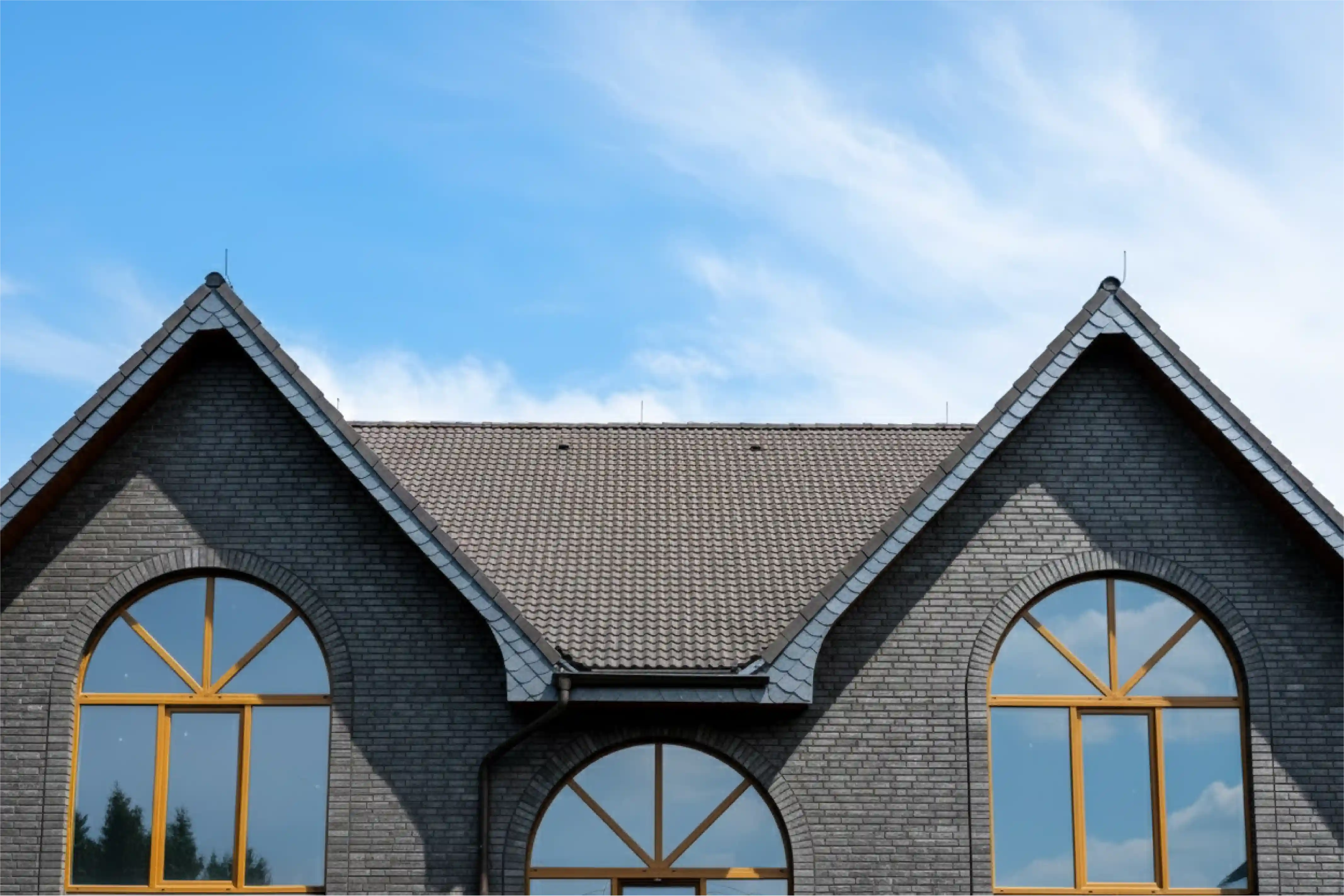 Close-up of a dark brick house roof with three large arched windows reflecting the blue sky.