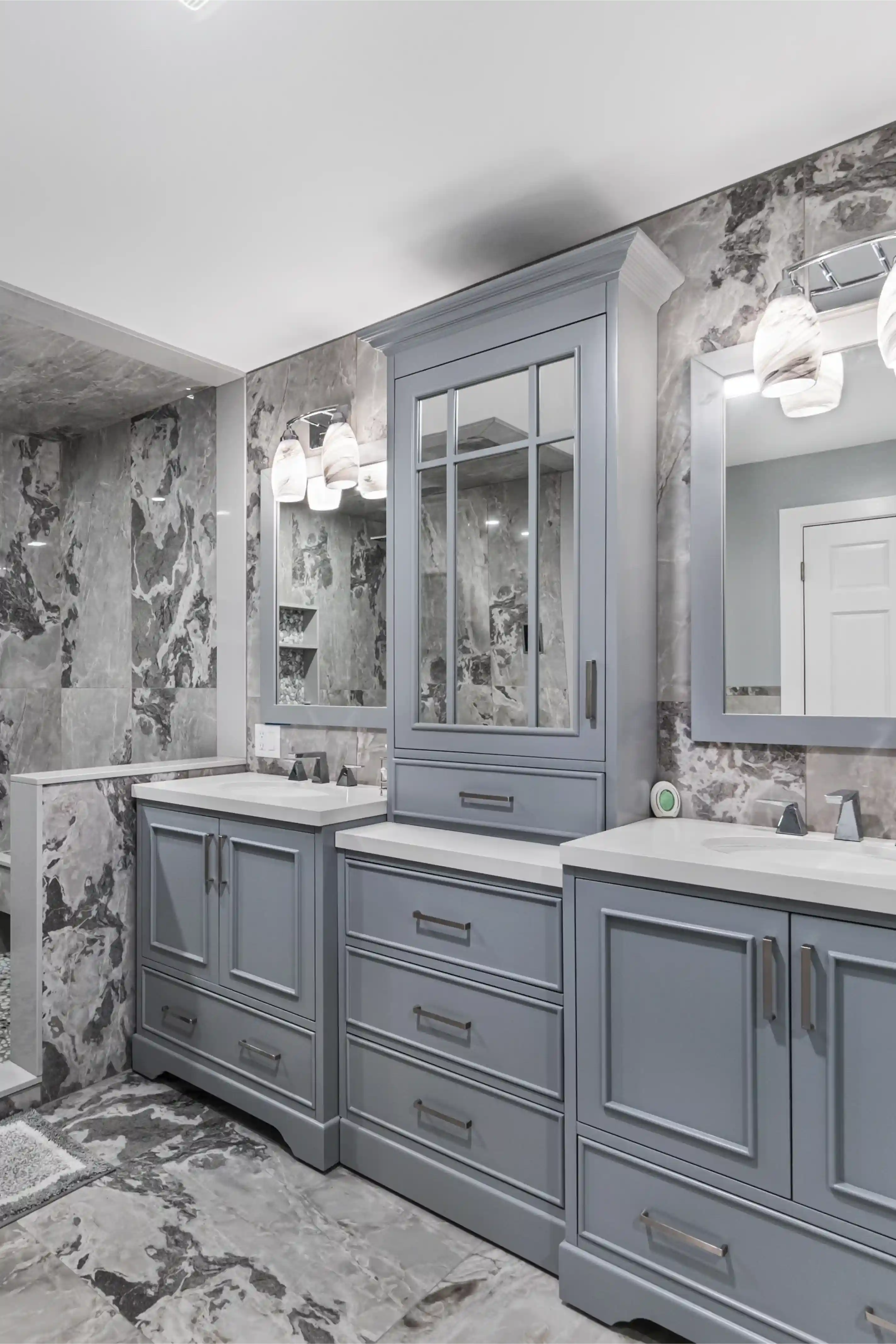 Modern bathroom with gray cabinetry, white countertops, dual sinks, and marble-patterned gray tiles on walls and floor.