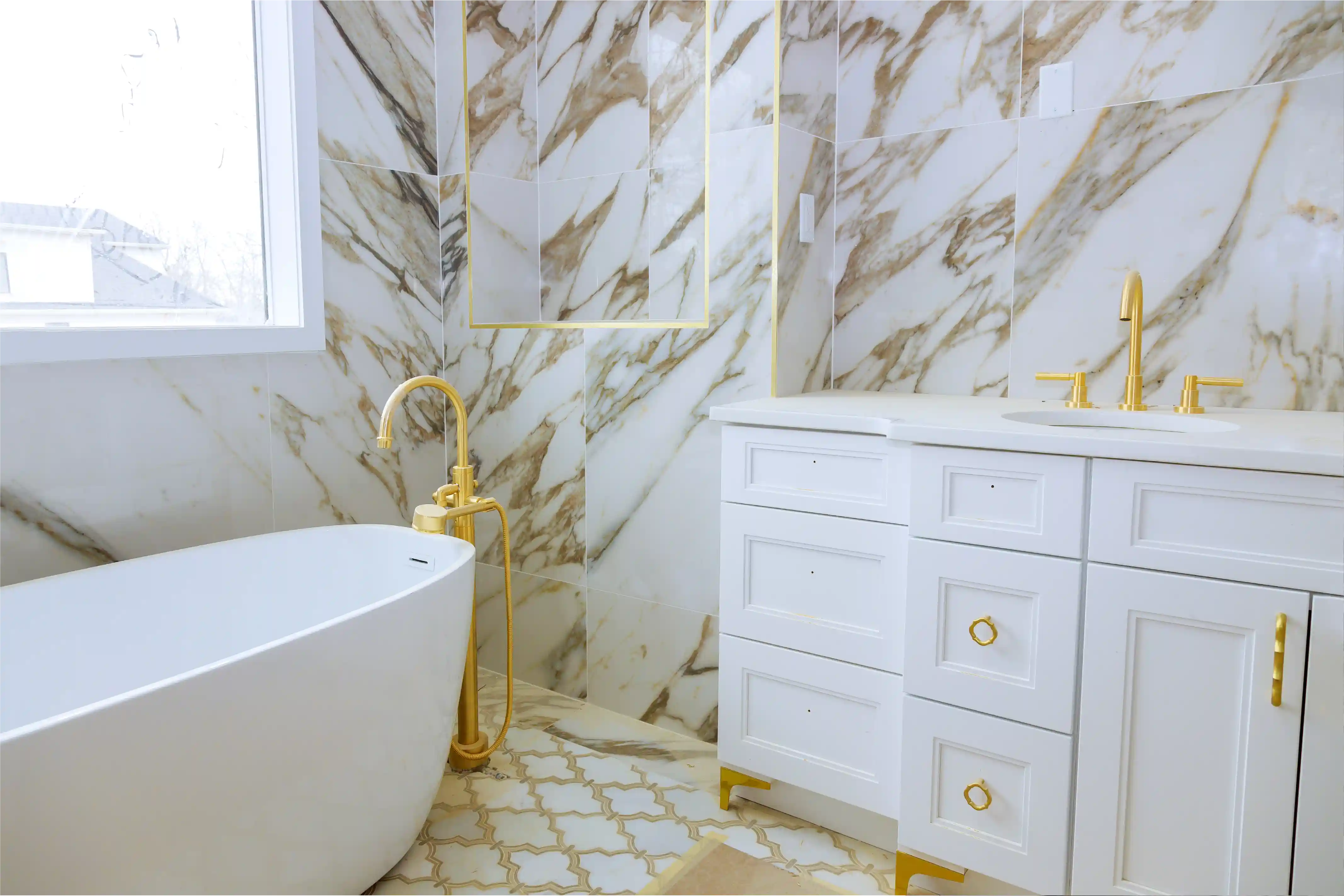 Modern bathroom with white freestanding bathtub, white vanity with gold fixtures, and marble-patterned walls and floor tiles.