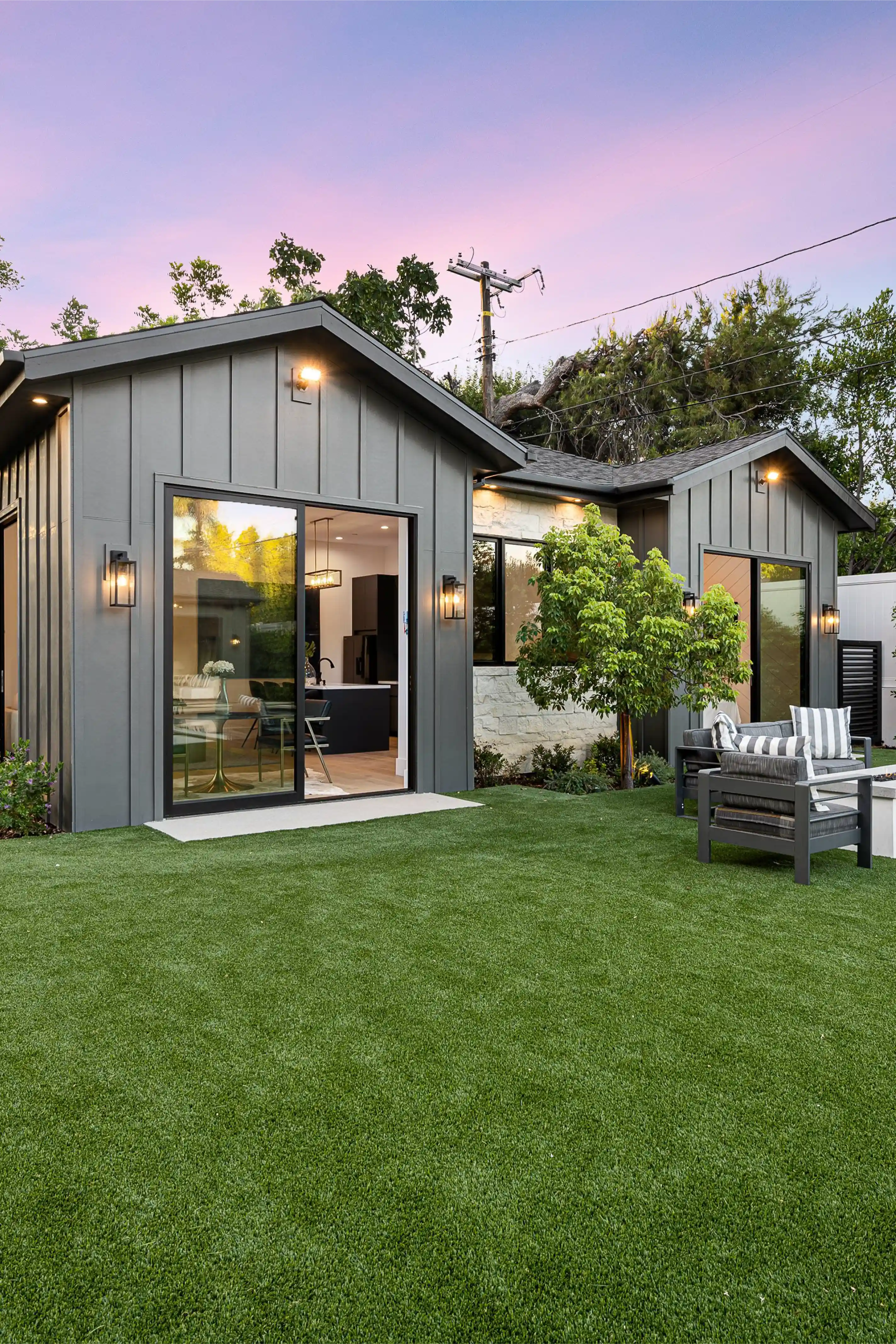 Modern gray ADU with large sliding glass doors, outdoor seating area with striped cushions, and lush green lawn at sunset.