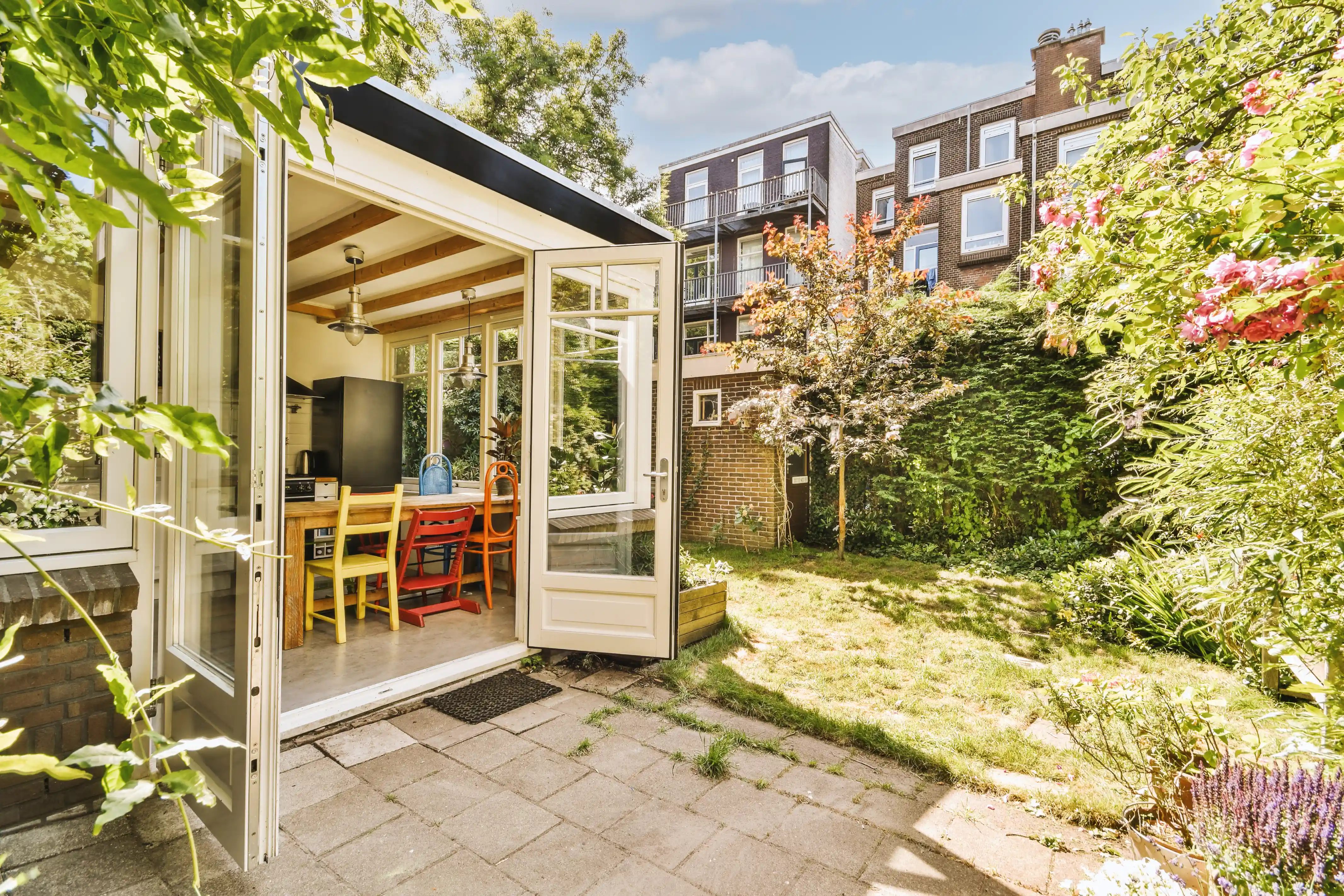 Open glass doors leading from a bright indoor dining area with colorful chairs to a sunlit backyard garden with grass, trees, and flowers.