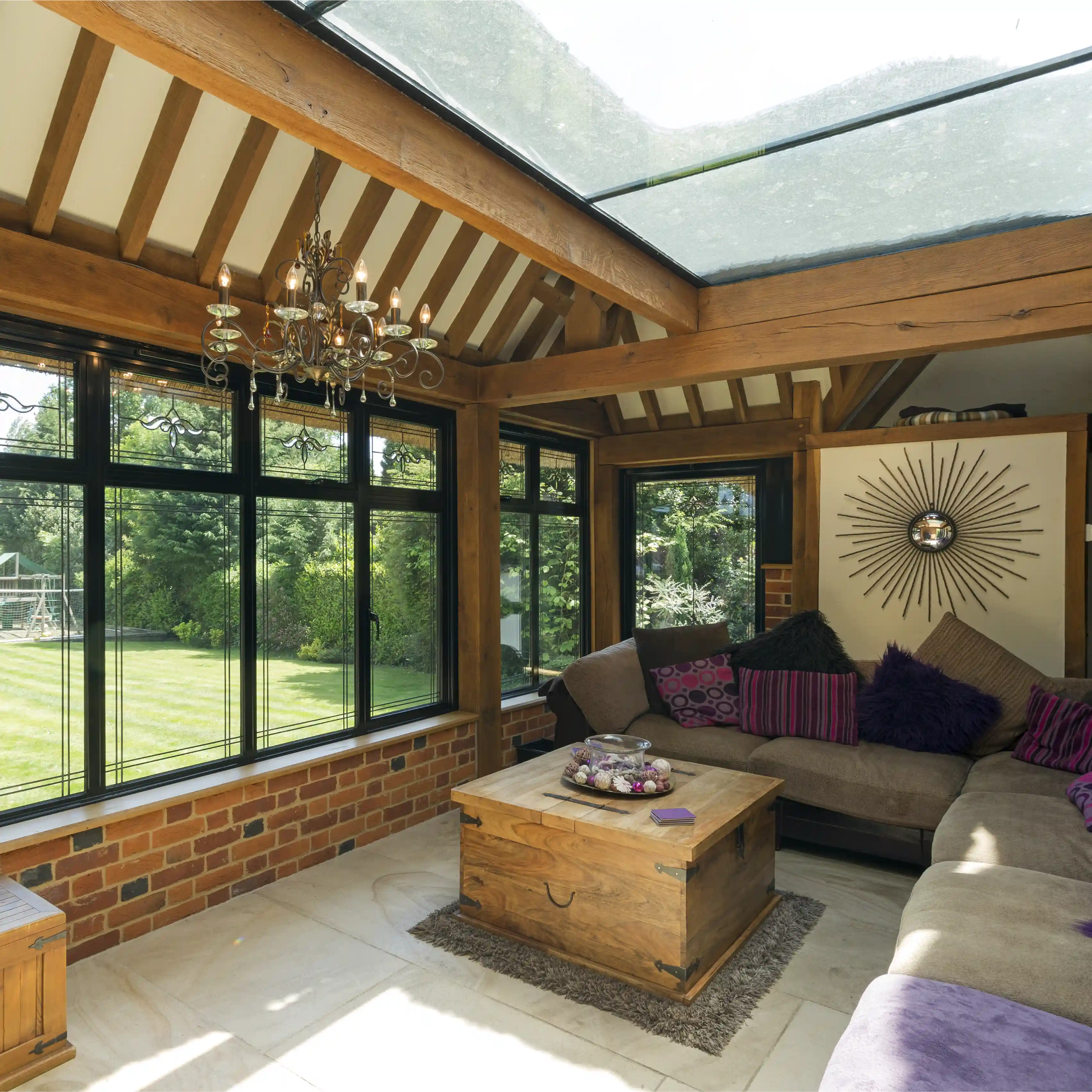 Sunroom with wooden beams, large black-framed windows overlooking a green lawn, a chandelier, and a sectional sofa with purple cushions around a wooden chest coffee table.