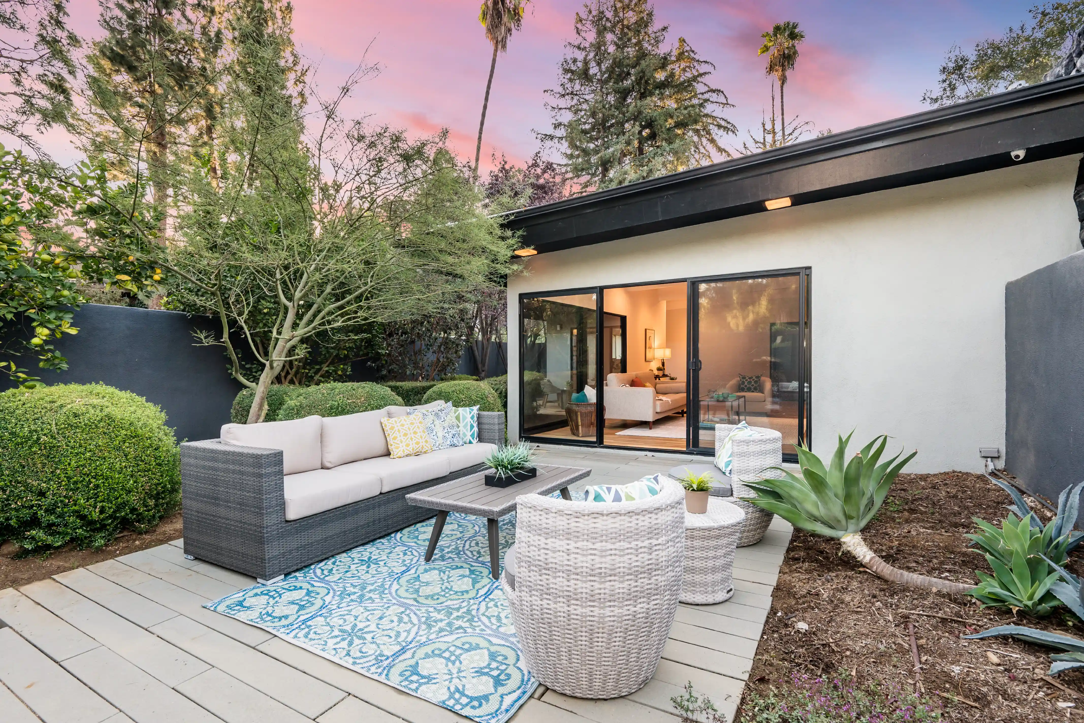 Outdoor patio with cushioned gray sofa, two white wicker chairs, and a patterned blue rug, surrounded by greenery and a sliding glass door to the interior.
