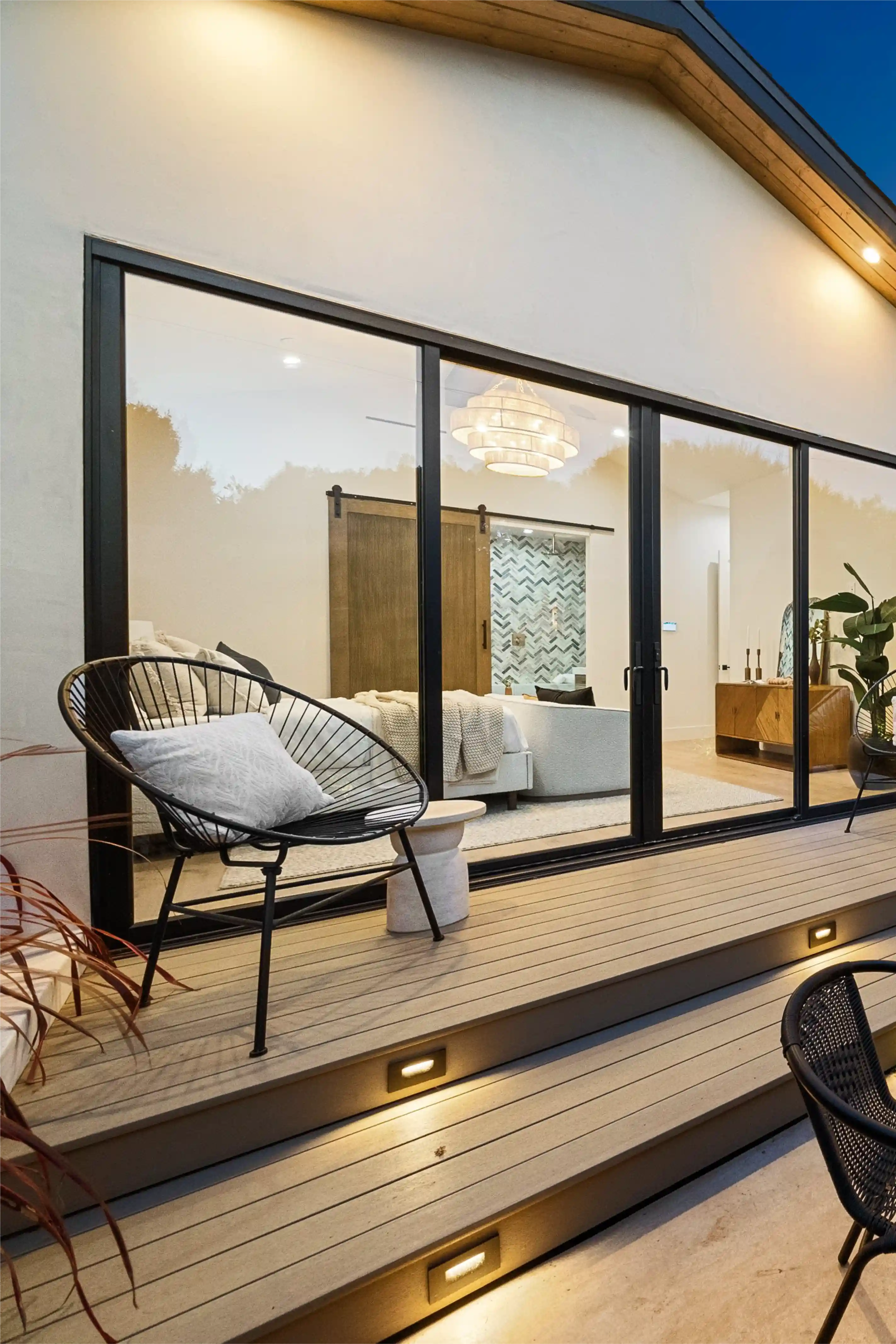 Modern patio with wooden steps lit by recessed lights, black metal chairs with white cushions, and a view through large glass doors into a bedroom with a wooden sliding door and elegant chandelier.