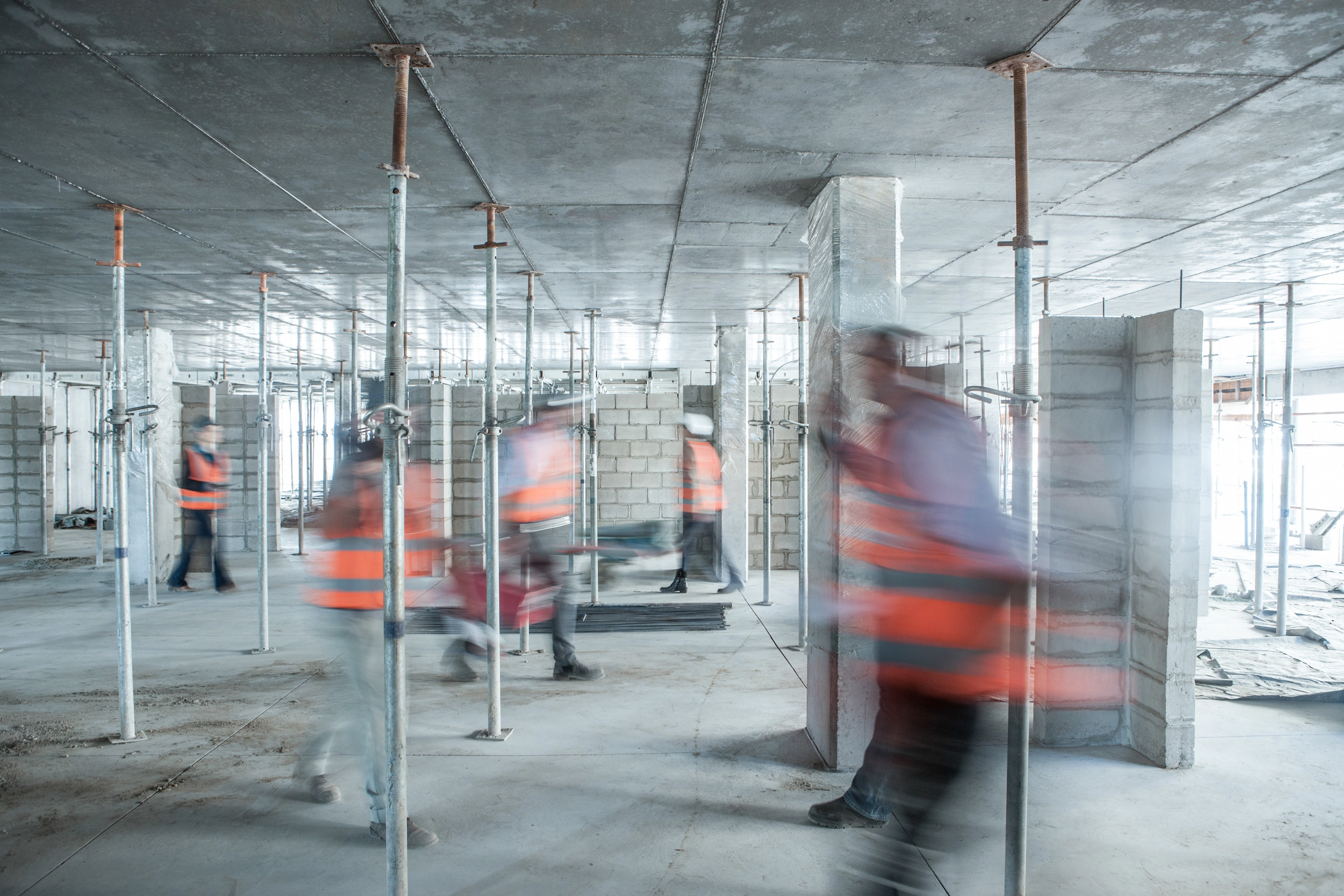 Construction workers in orange vests moving inside a large building under construction with metal supports.