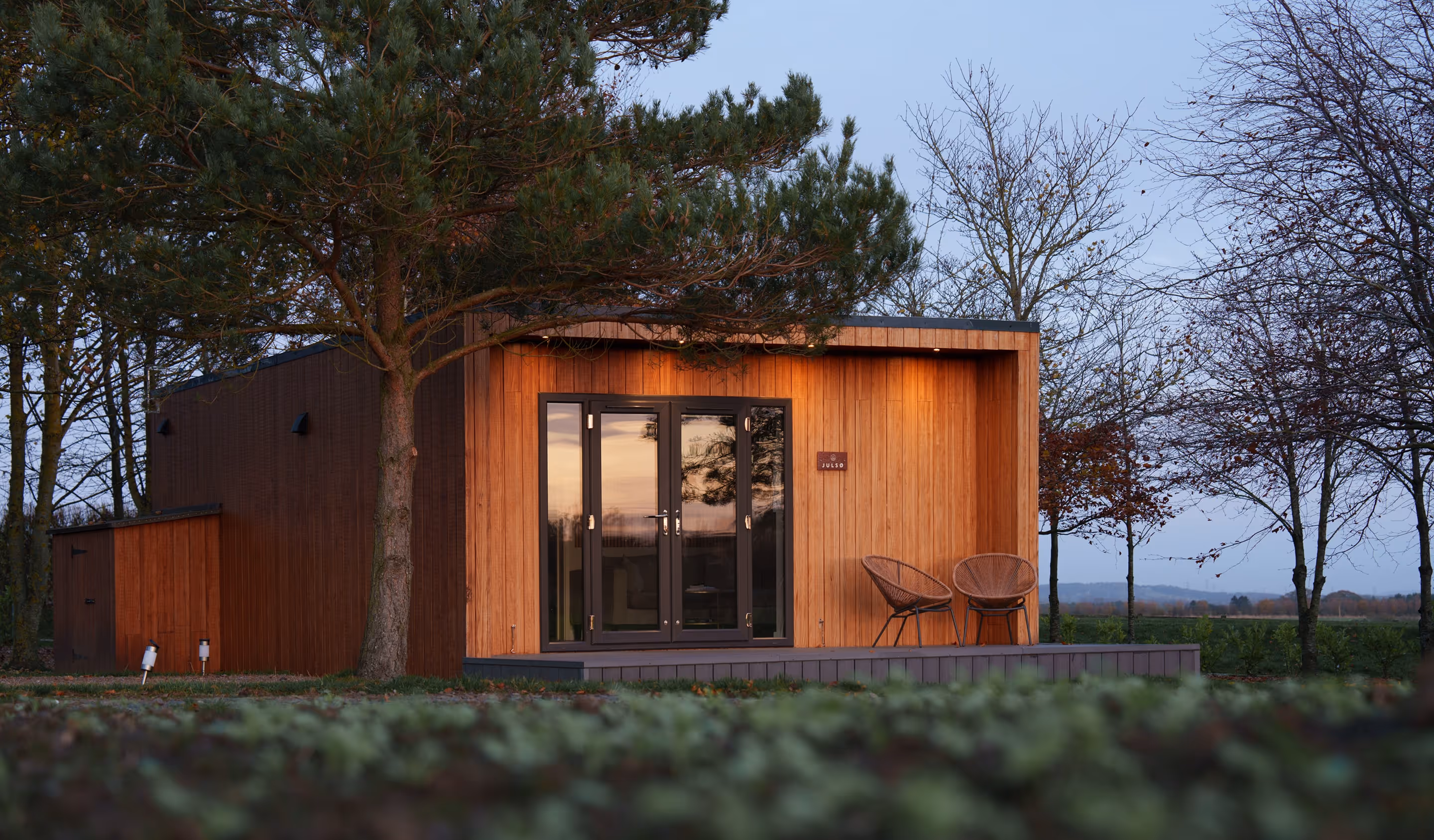 Modern wooden cabin with glass double doors and two chairs on the porch, surrounded by trees at dusk.
