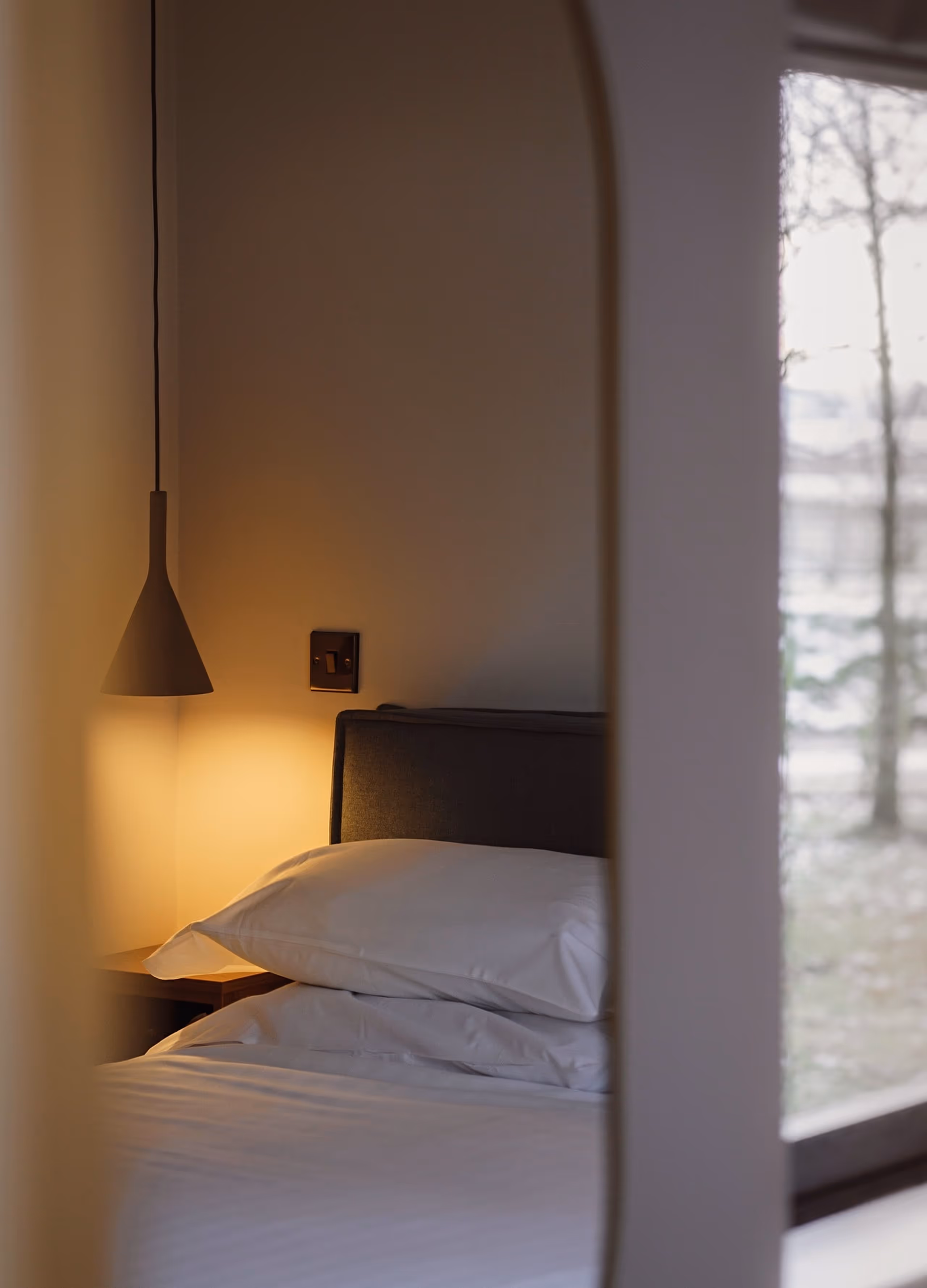Cozy bedroom corner with a bed, white pillows, a hanging lamp, and a window showing an outdoor view.