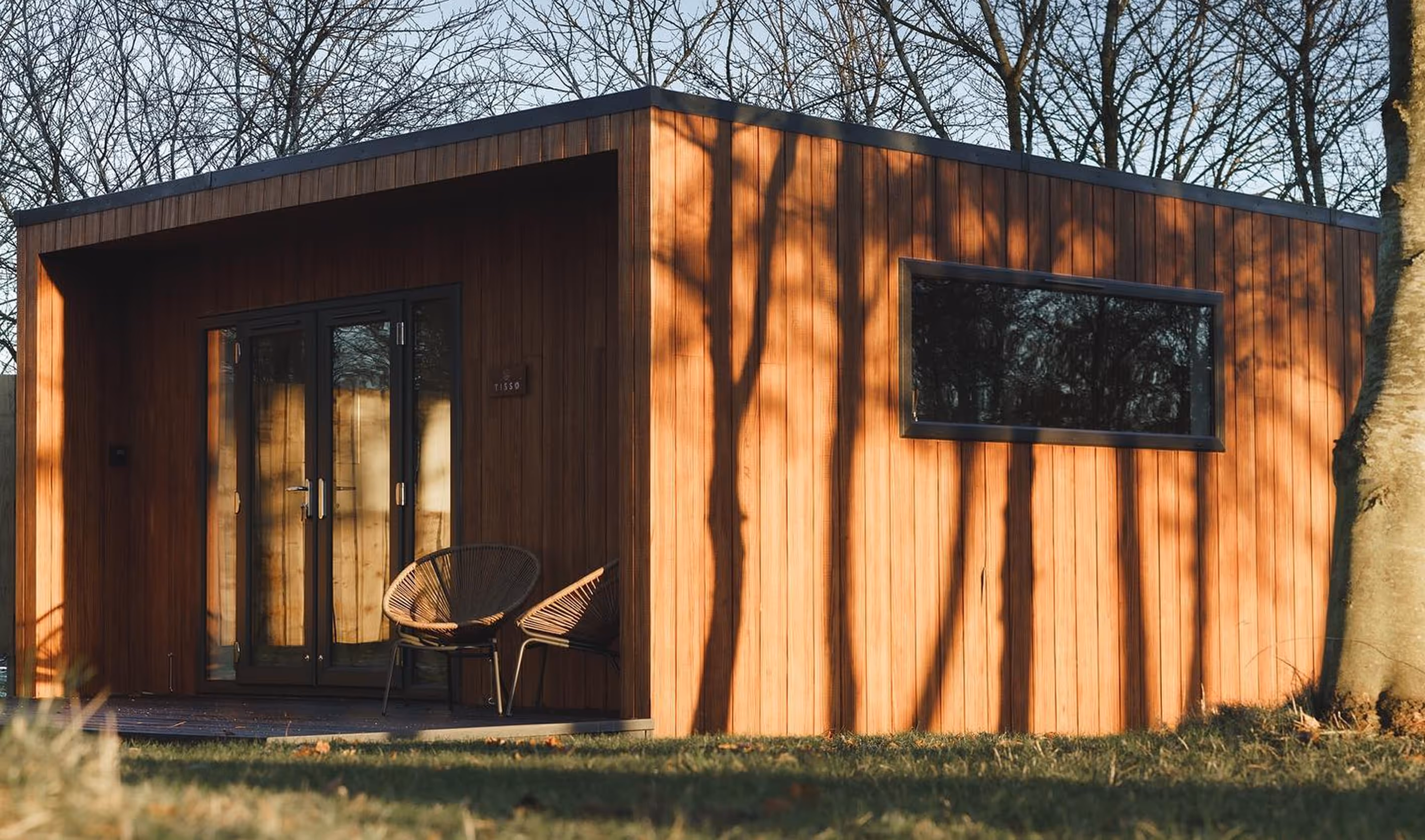 Modern wooden cabin with two chairs on a small porch, shadows of trees cast on its facade in a grassy area.
