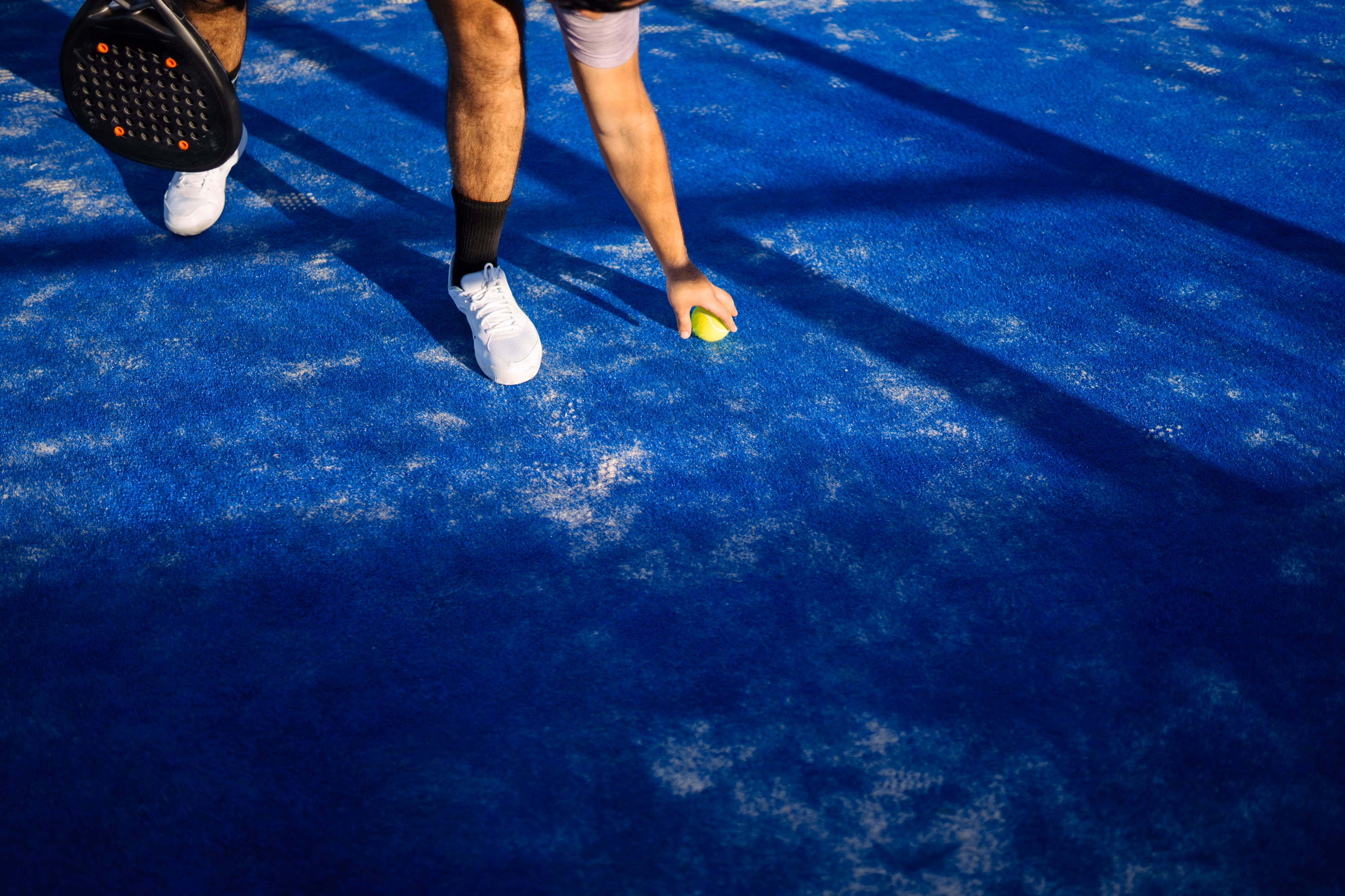 Player wearing white shoes and black socks reaches down to pick up a tennis ball on a blue tennis court holding a paddle in the other hand.