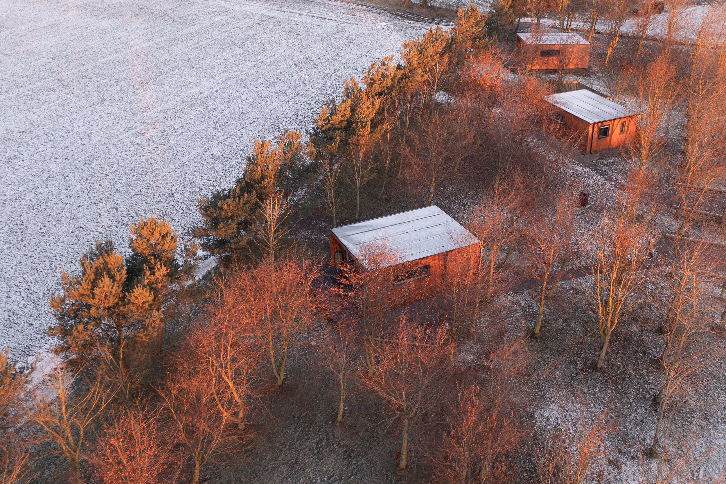Aerial view of small wooden cabins surrounded by leafless trees with a light dusting of snow and warm sunlight.