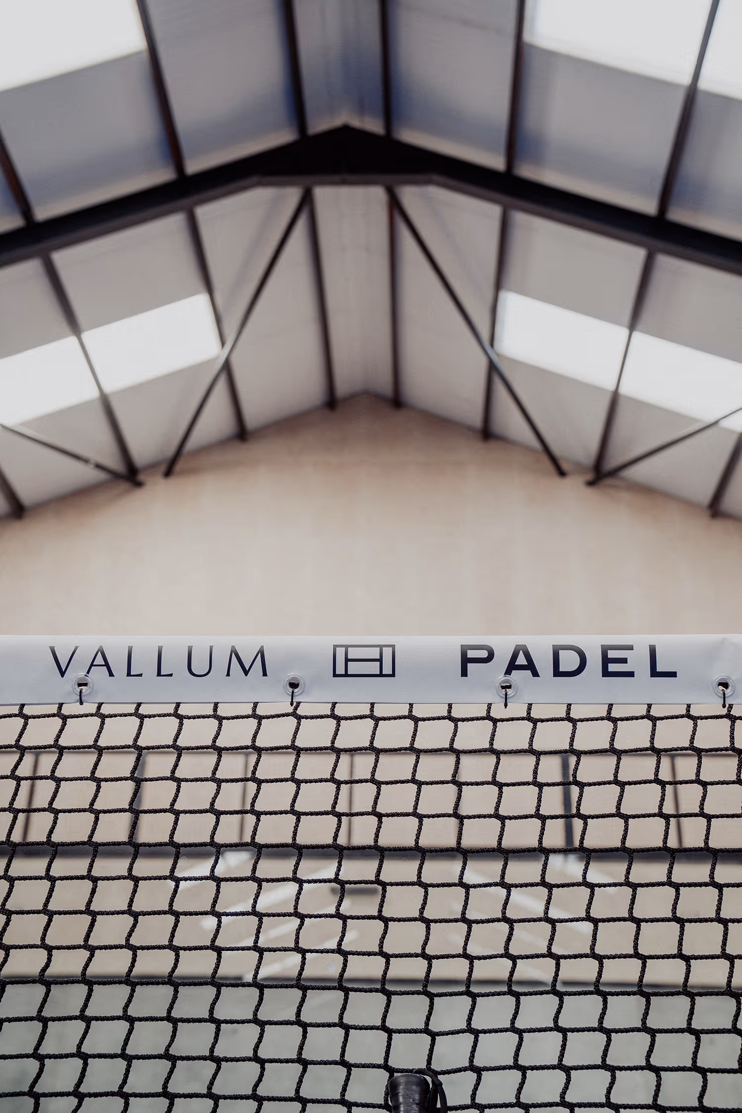 Black padel net with white band labeled Vallum Padel inside indoor court under a high ceiling with skylights.