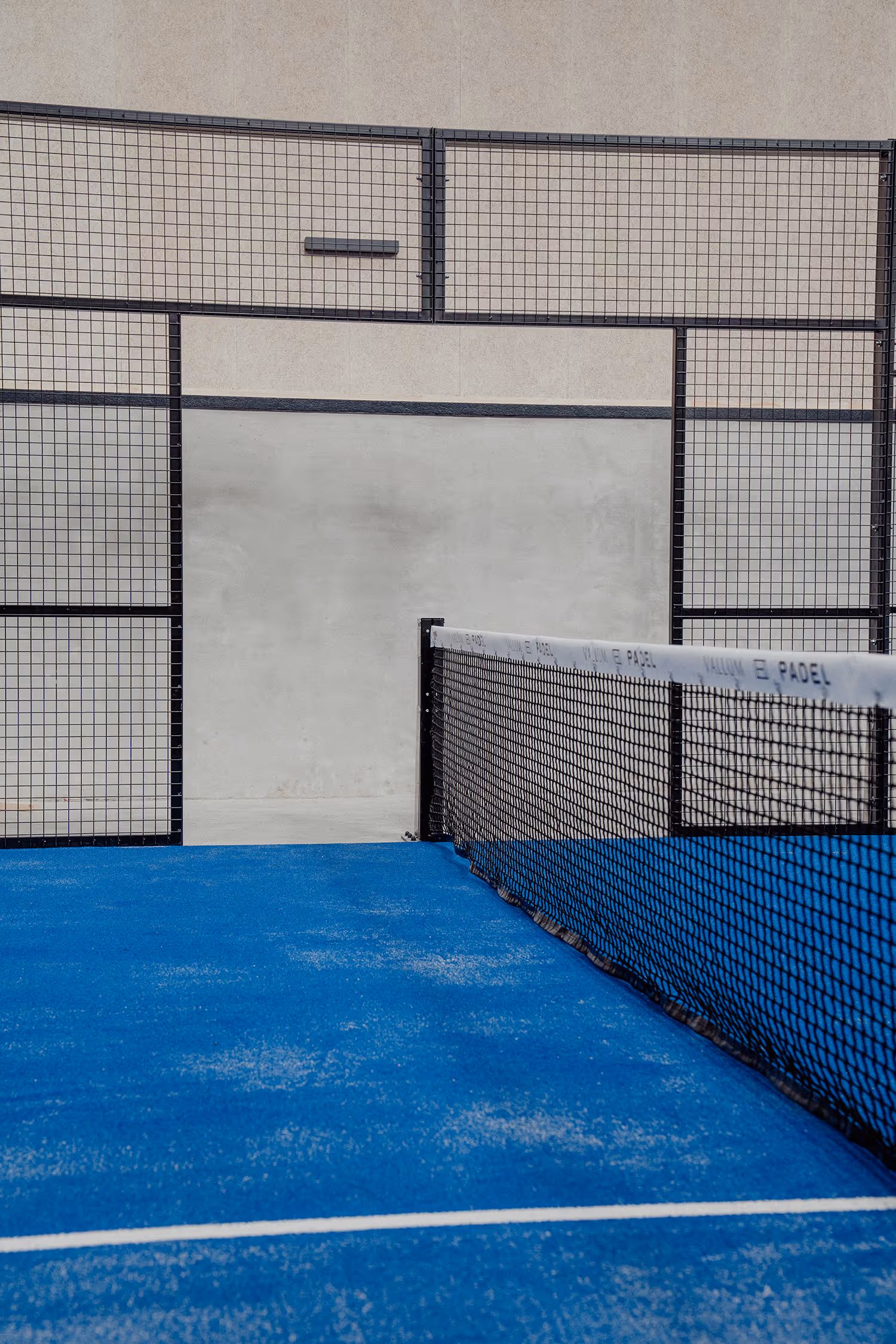 Close-up view of a blue padel court with a black net and a solid wall at the end.