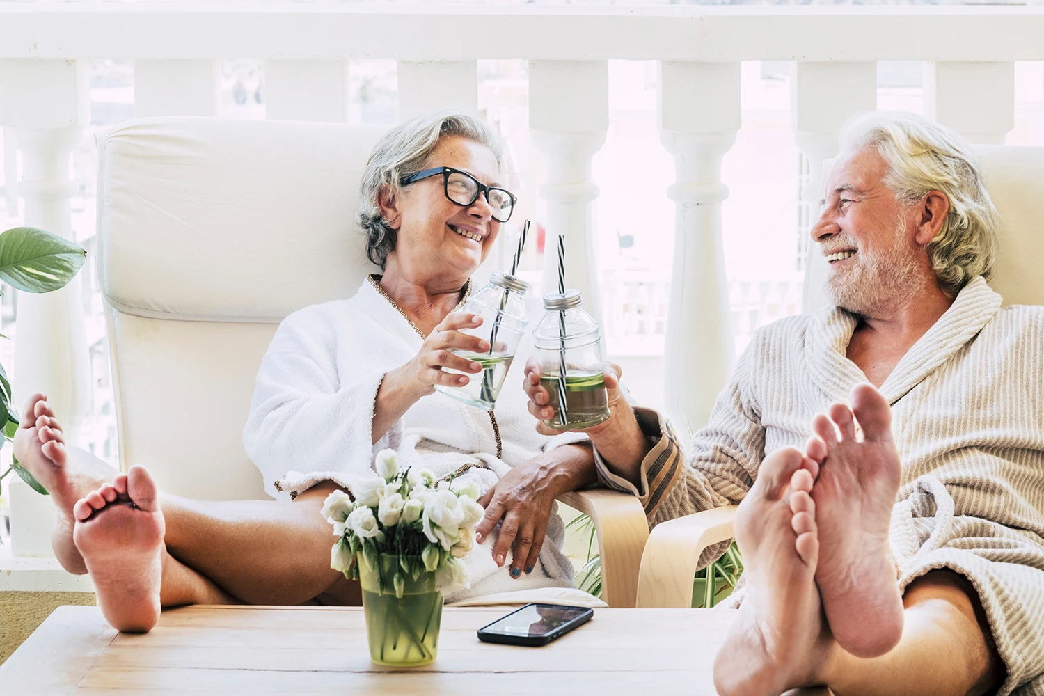 Smiling elderly couple in bathrobes relaxing on lounge chairs, toasting with drinks in glass jars.