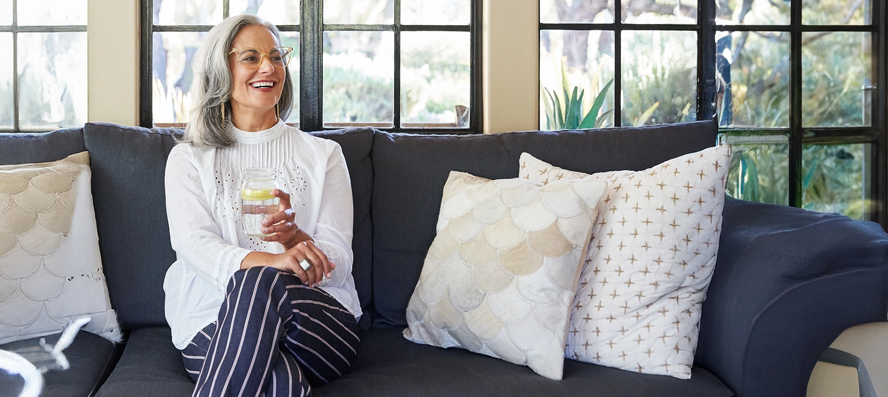 Smiling mature woman with glasses sitting on a couch holding a glass of water with lemon.