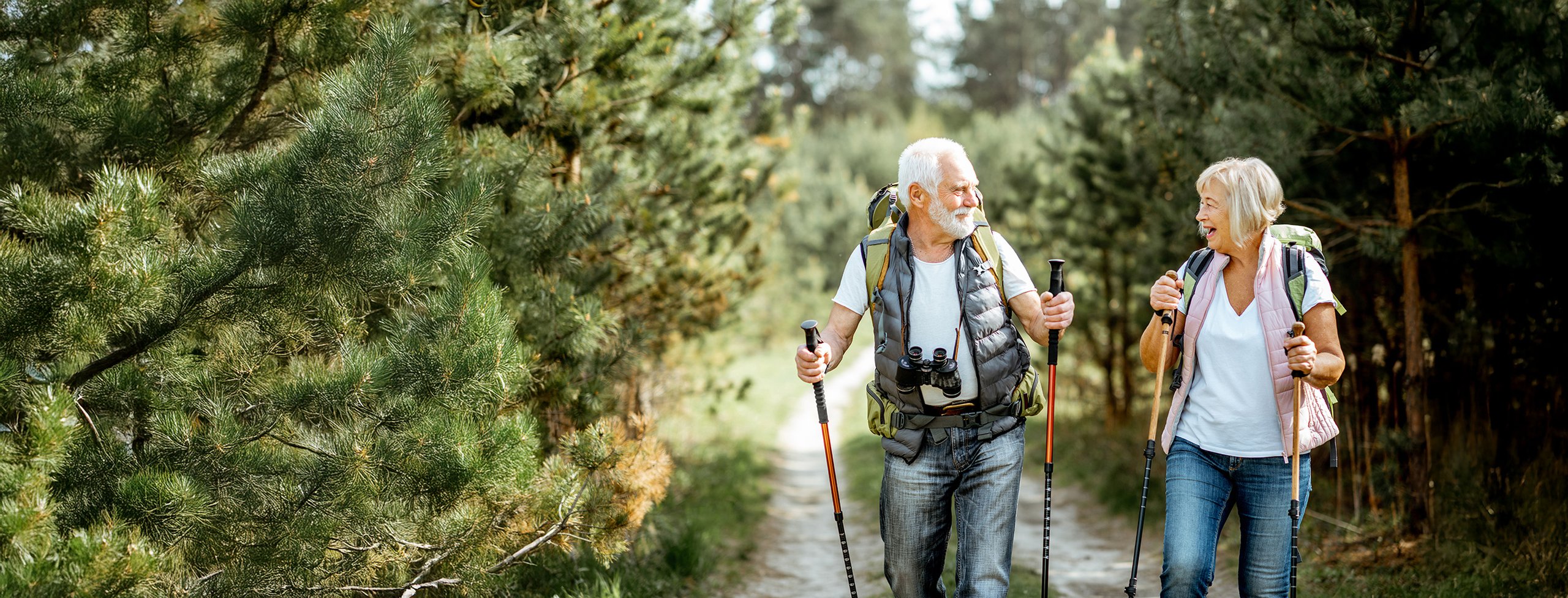 A senior man and woman hiking with backpacks and walking sticks on a forest trail, smiling at each other.