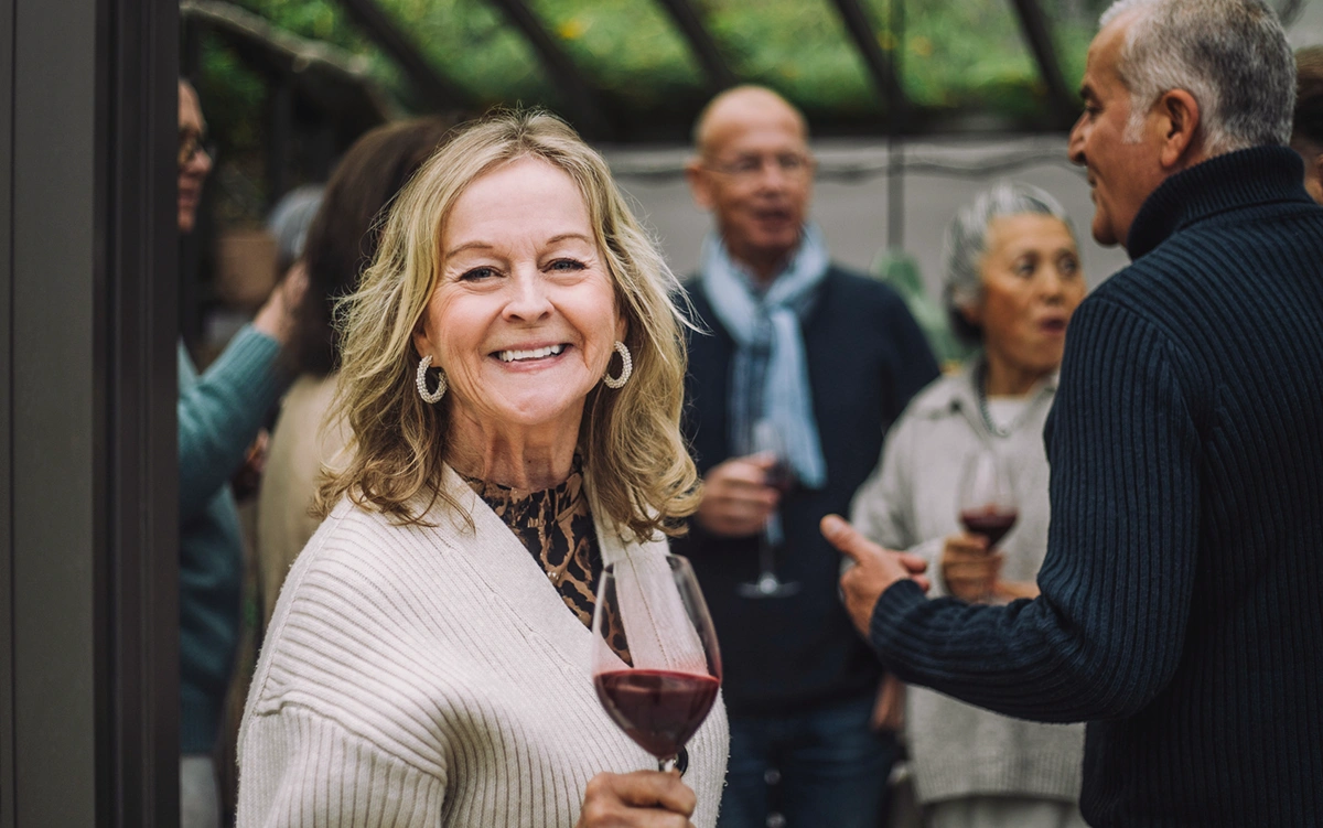Smiling woman holding a glass of red wine at a social gathering with people conversing in the background.