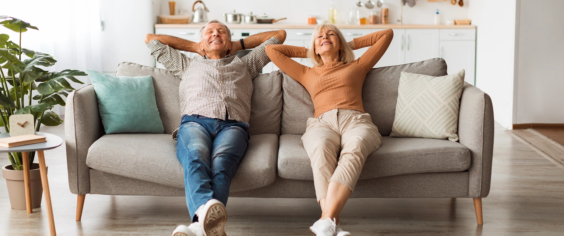 Older man and woman reclining with hands behind their heads on a gray sofa in a bright living room.