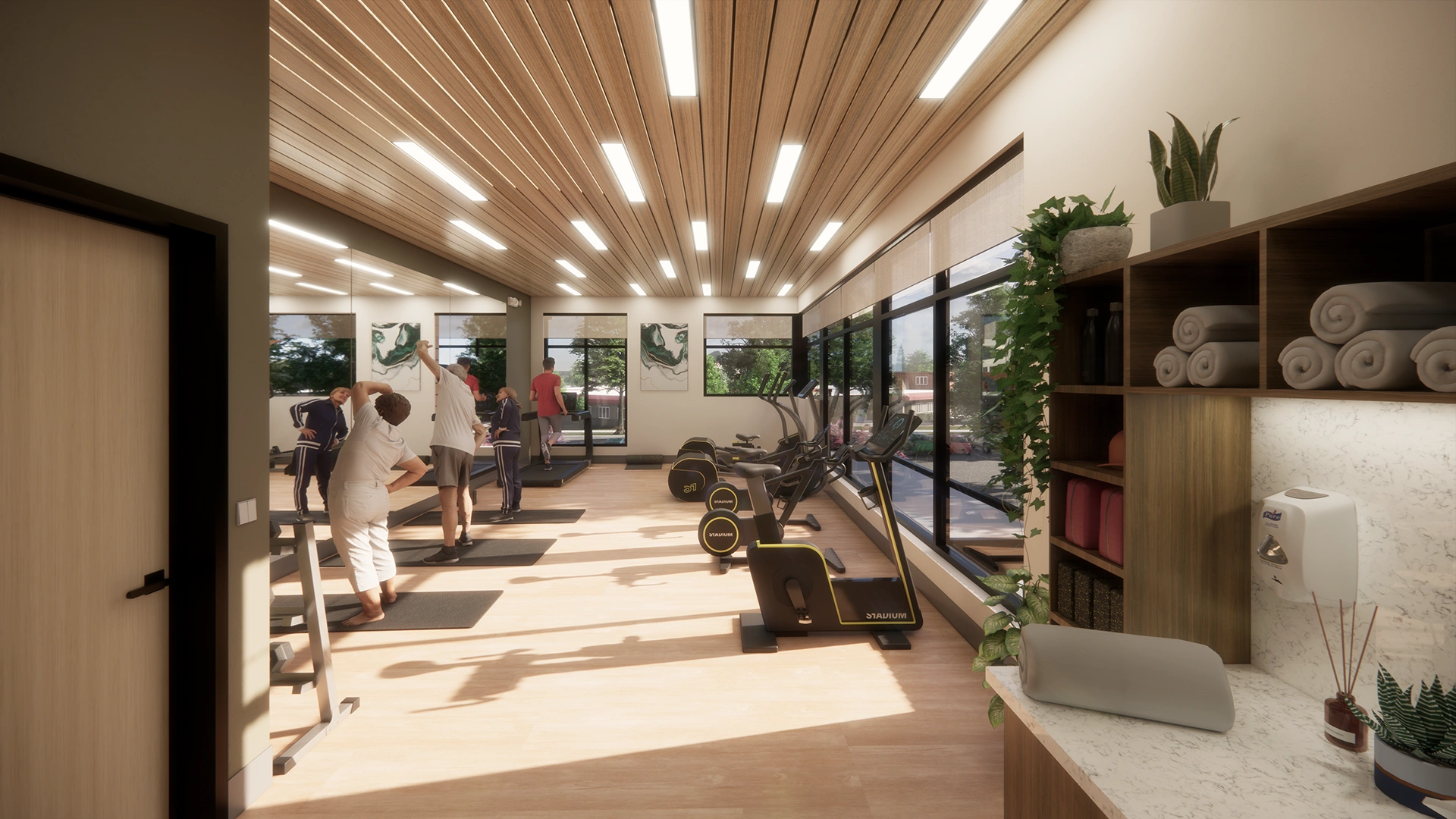 Bright fitness center with large windows, exercise bikes, treadmills, people stretching on mats, and shelves with rolled towels and plants.
