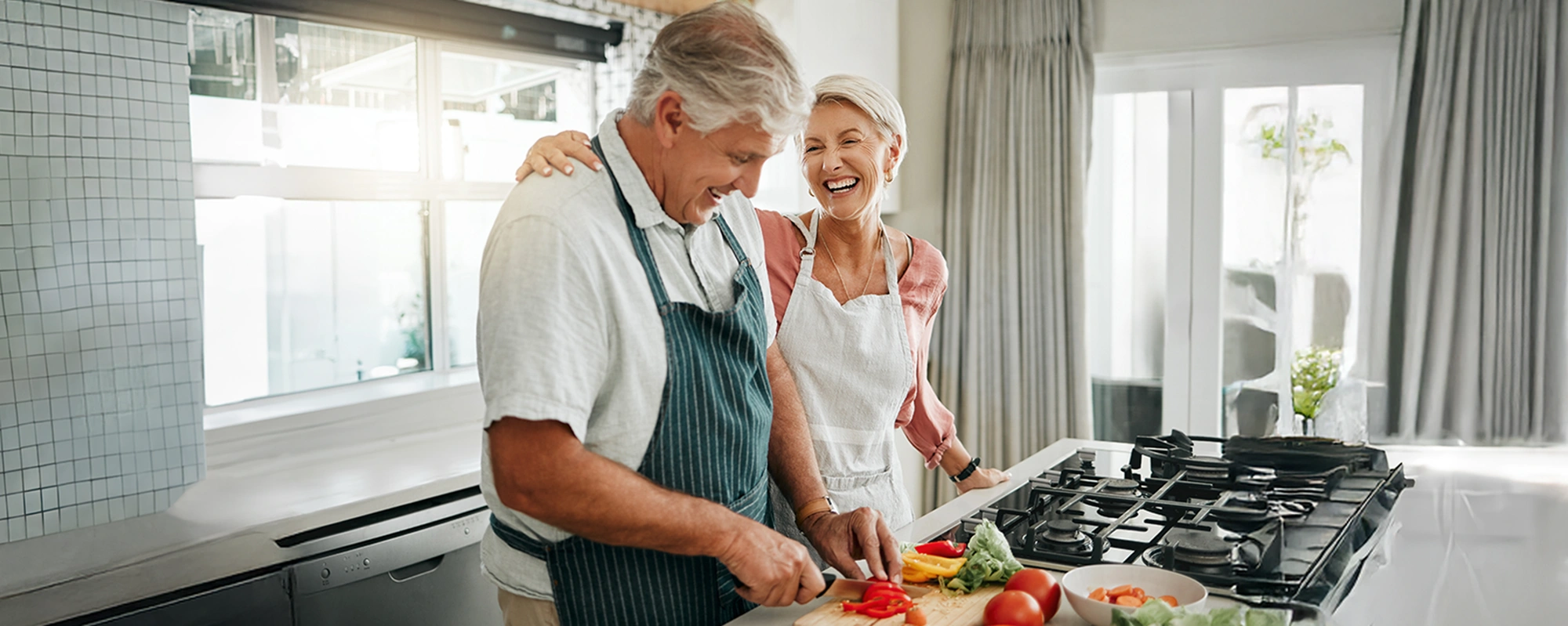 Senior couple wearing aprons laughing and preparing vegetables together in a bright kitchen.