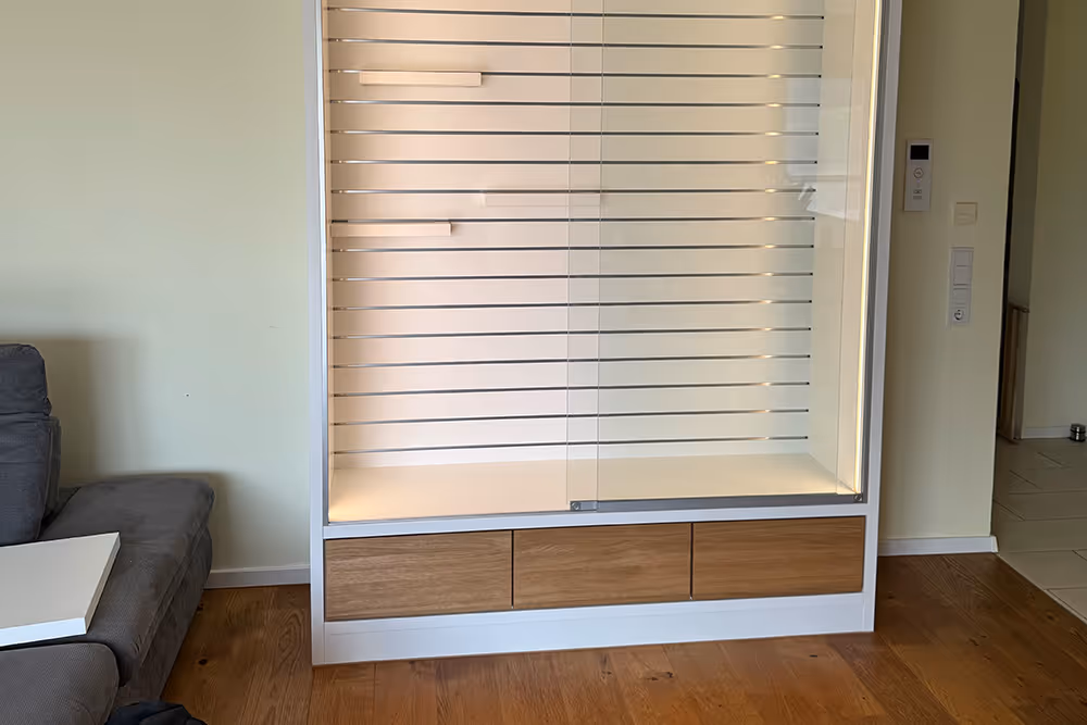 Leeres, weißes Vitrinenregal mit Holzschubladen unten in einem Raum mit Holzboden neben einem grauen Sofa.