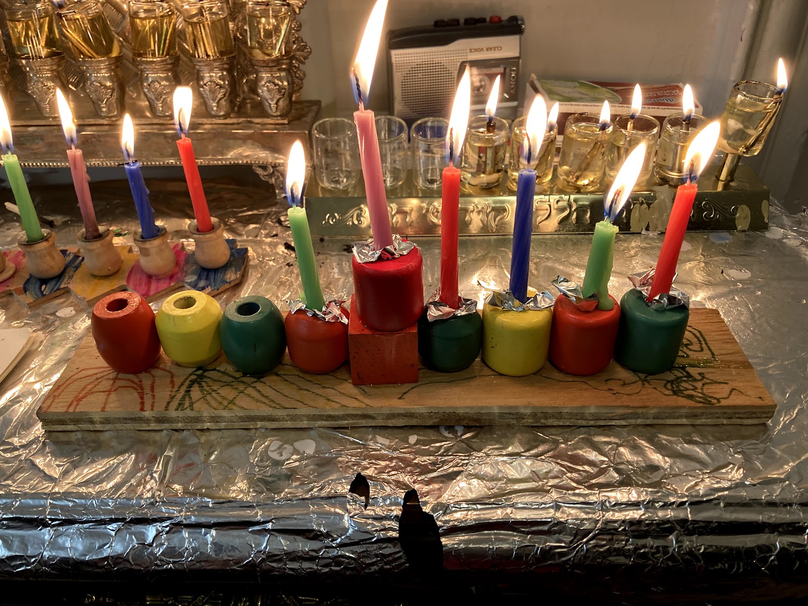 Lit Hanukkah menorah with colorful candles on a wooden base covered in foil, with another menorah and dreidels in the background.