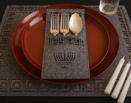 A grey felt placemat with a menorah cutlery pocket on a red charger, with candles ready to light