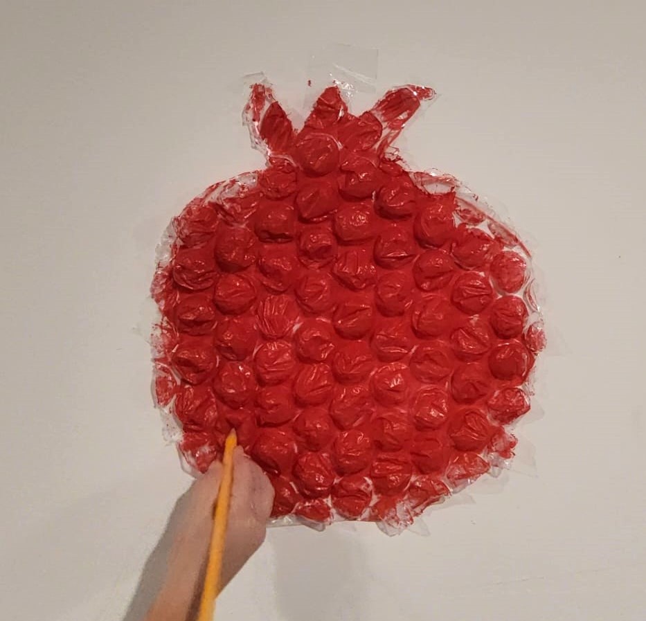A sheet of large bubble wrap cut into a pomegranate shape and painted red, taped to a wall, with a child holding a paintbrush up to it.