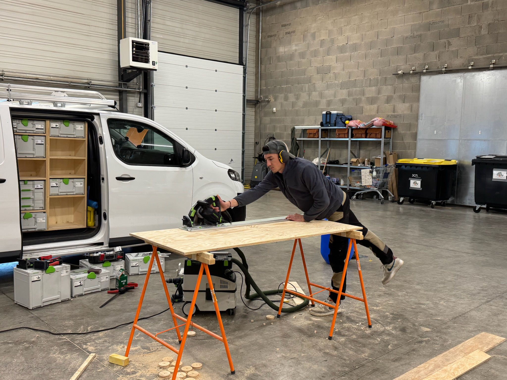 Un ouvrier en équipements de protection utilise une scie circulaire pour couper une plaque de bois sur un établi dans un atelier.