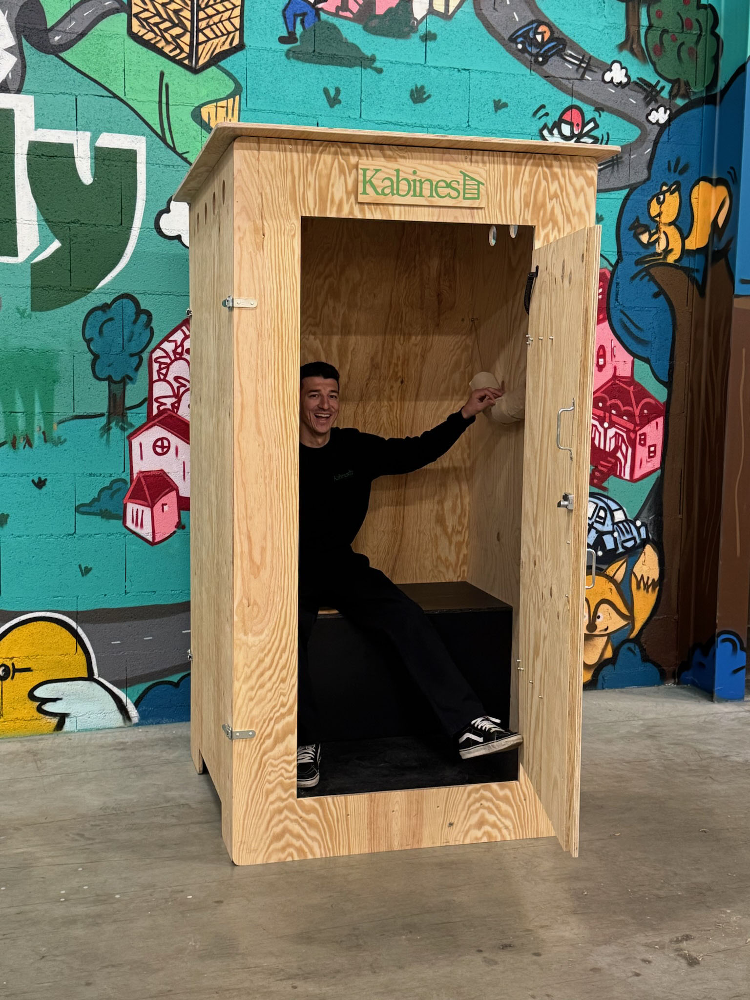 Un homme souriant est assis à l'intérieur d'une cabine en bois ouverte marquée 'Kabines' devant un mur peint coloré.