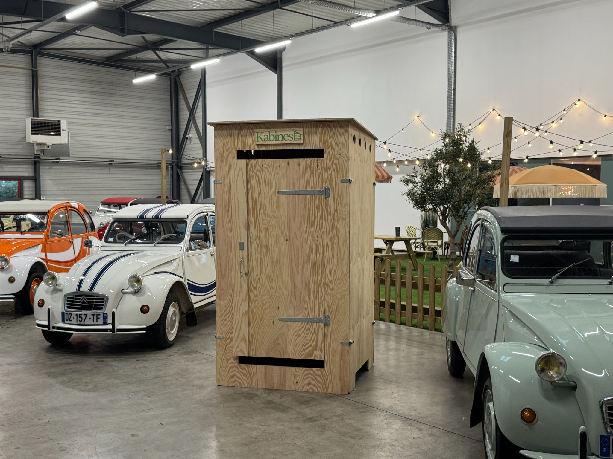Cabine en bois avec le signe Kabines au centre d'un garage abritant plusieurs voitures Citroën 2CV colorées.