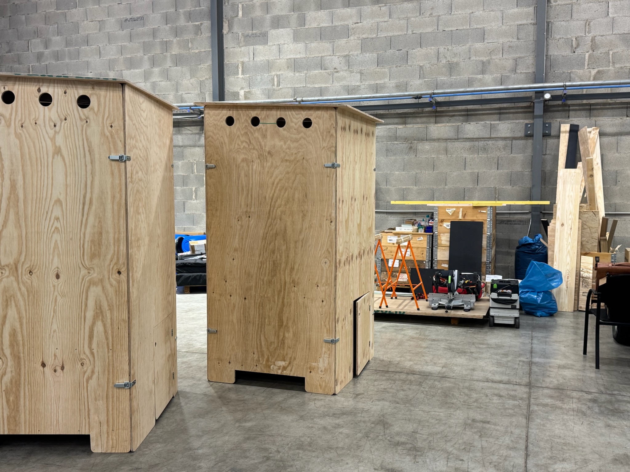 Deux cabines en bois brut avec des trous ronds en haut, placés dans un atelier avec des outils et équipements en arrière-plan.