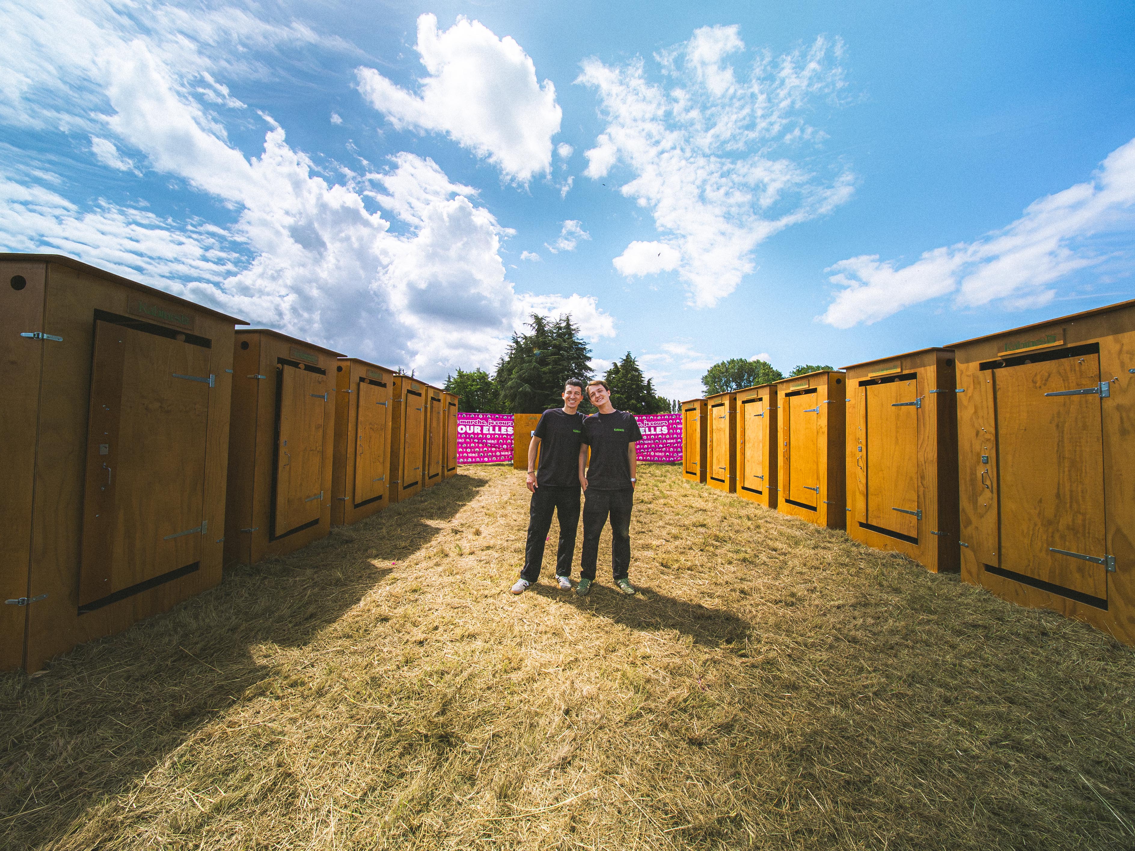 Deux jeunes hommes debout entre rangées de toilettes en bois sous un ciel partiellement nuageux.