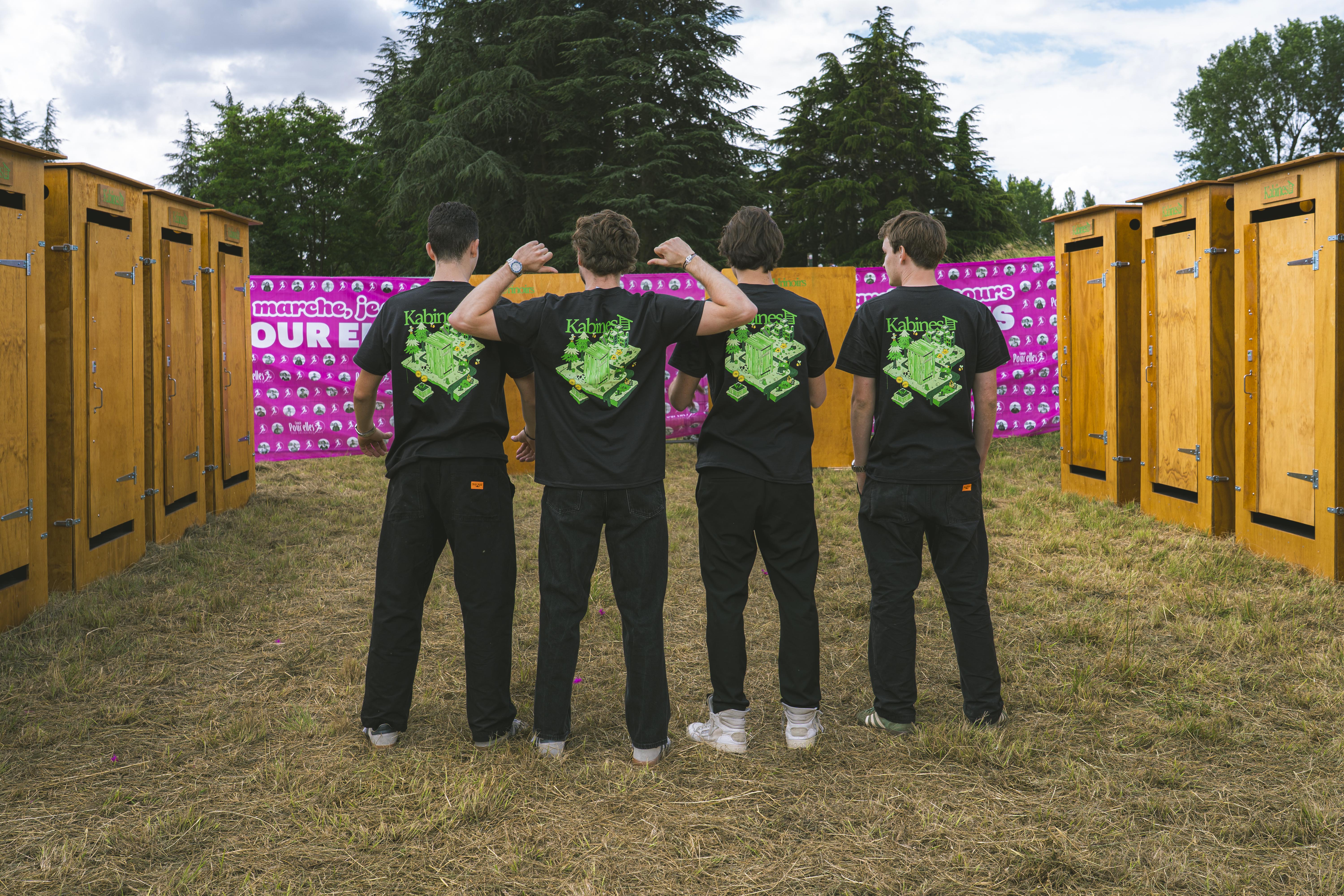 Quatre jeunes hommes avec des t-shirts noirs portant un design vert, debout devant une rangée de cabines en bois.