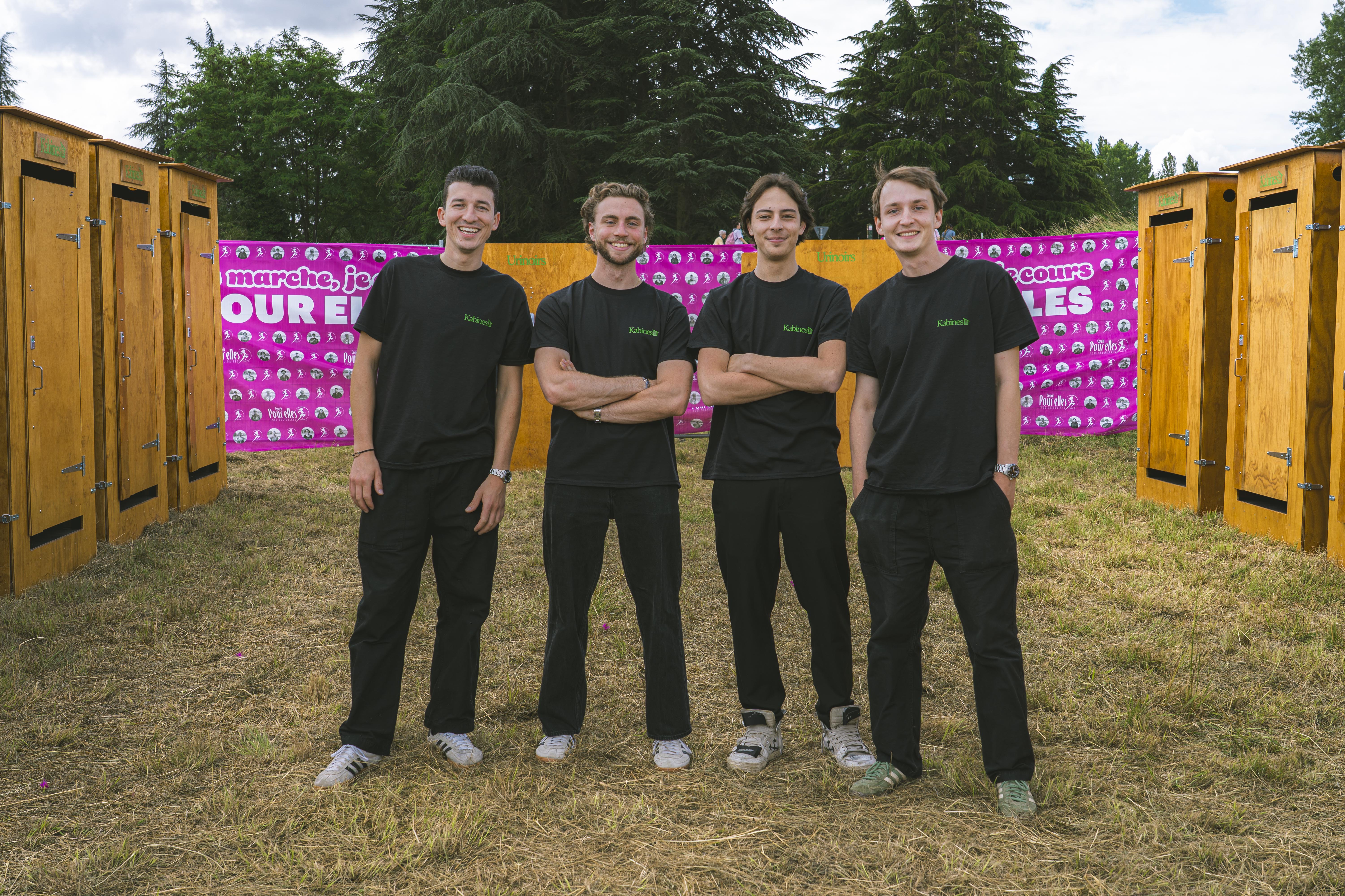 Quatre jeunes hommes souriants en t-shirt noir debout devant des toilettes portables en bois.