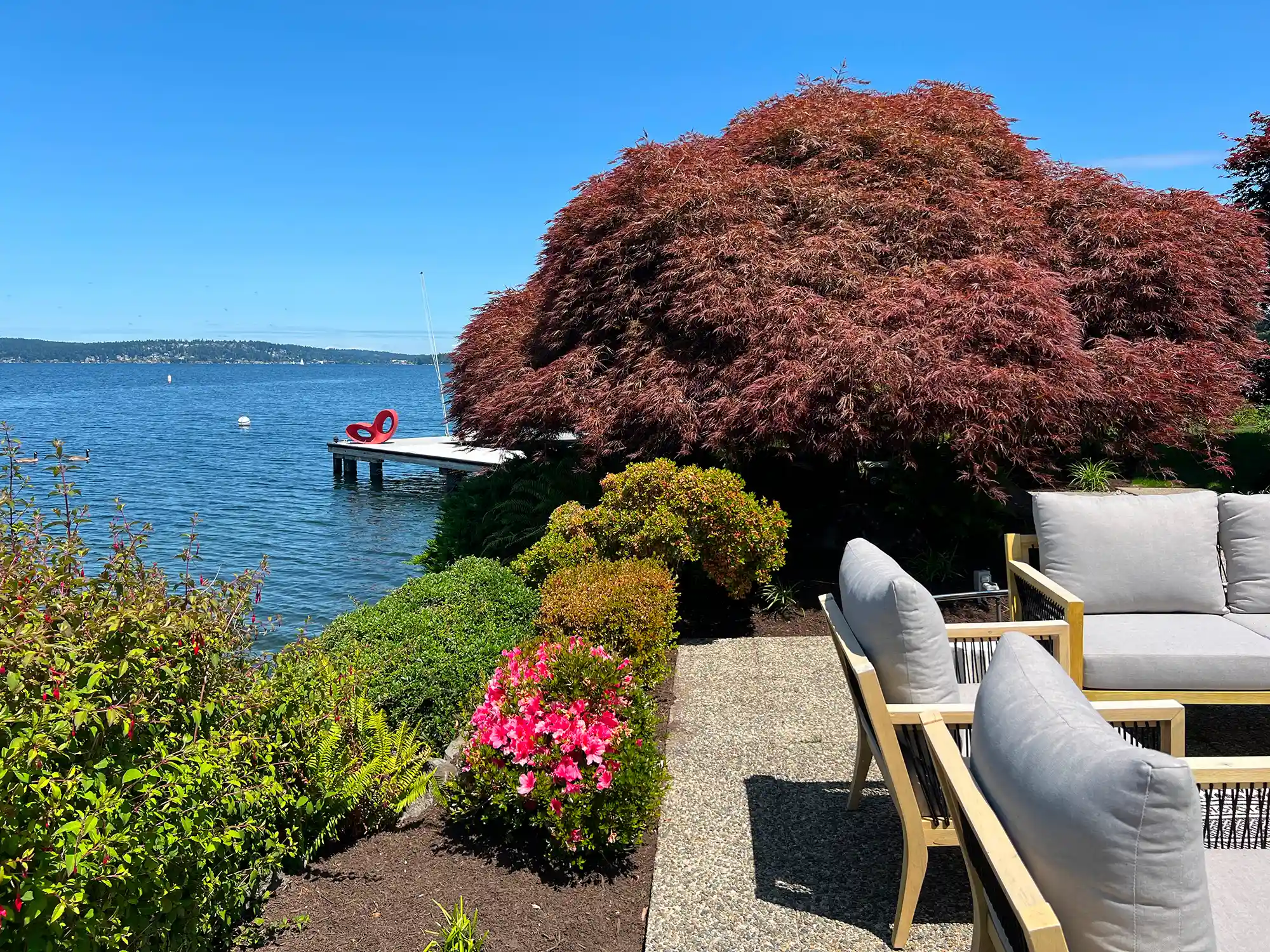 Patio with light gray cushioned chairs next to a garden with green and red bushes overlooking a lake with a wooden dock and a red chair.