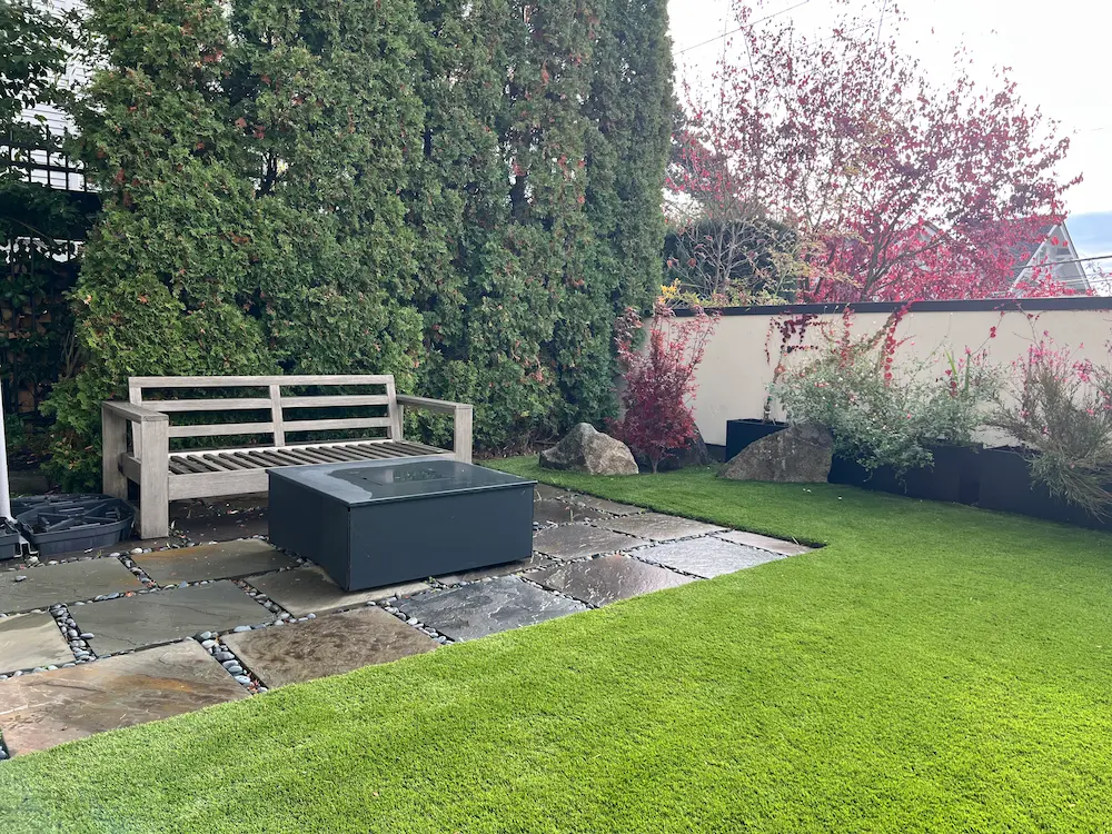 Outdoor patio with a wooden bench, a square black fire pit, stone tiles, and lush green lawn surrounded by tall evergreen trees and flowering shrubs.