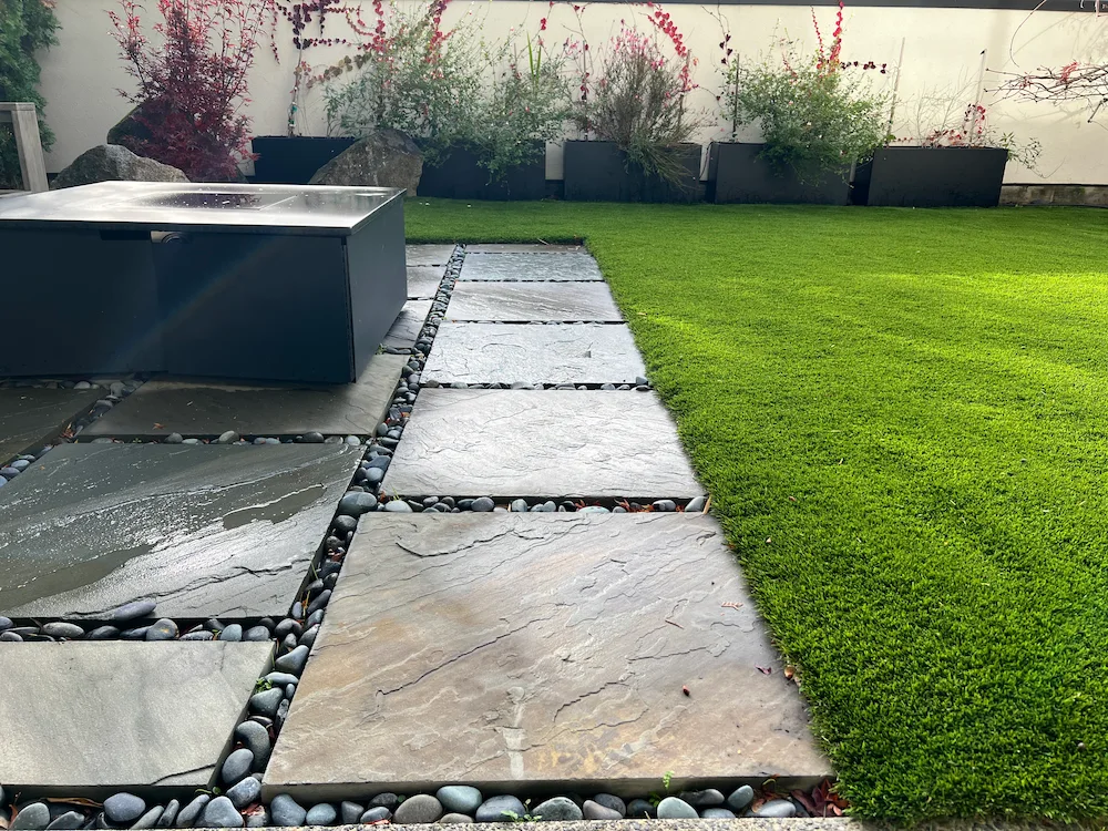 Outdoor garden with a pathway of stone slabs bordered by gray pebbles leading past a modern black metal fire pit to a green lawn and potted shrubs against a white wall.