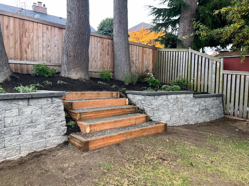 Wooden steps bordered by gray stone retaining walls leading up to a garden bed with trees and plants, next to a wooden fence.