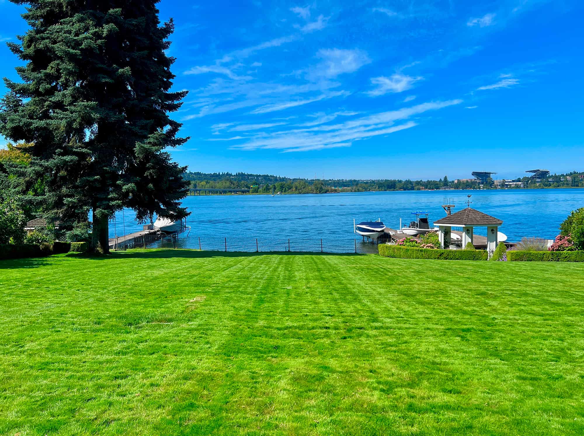 Well-maintained green lawn with a large tree on the left, overlooking a lake with boats docked and a clear blue sky.